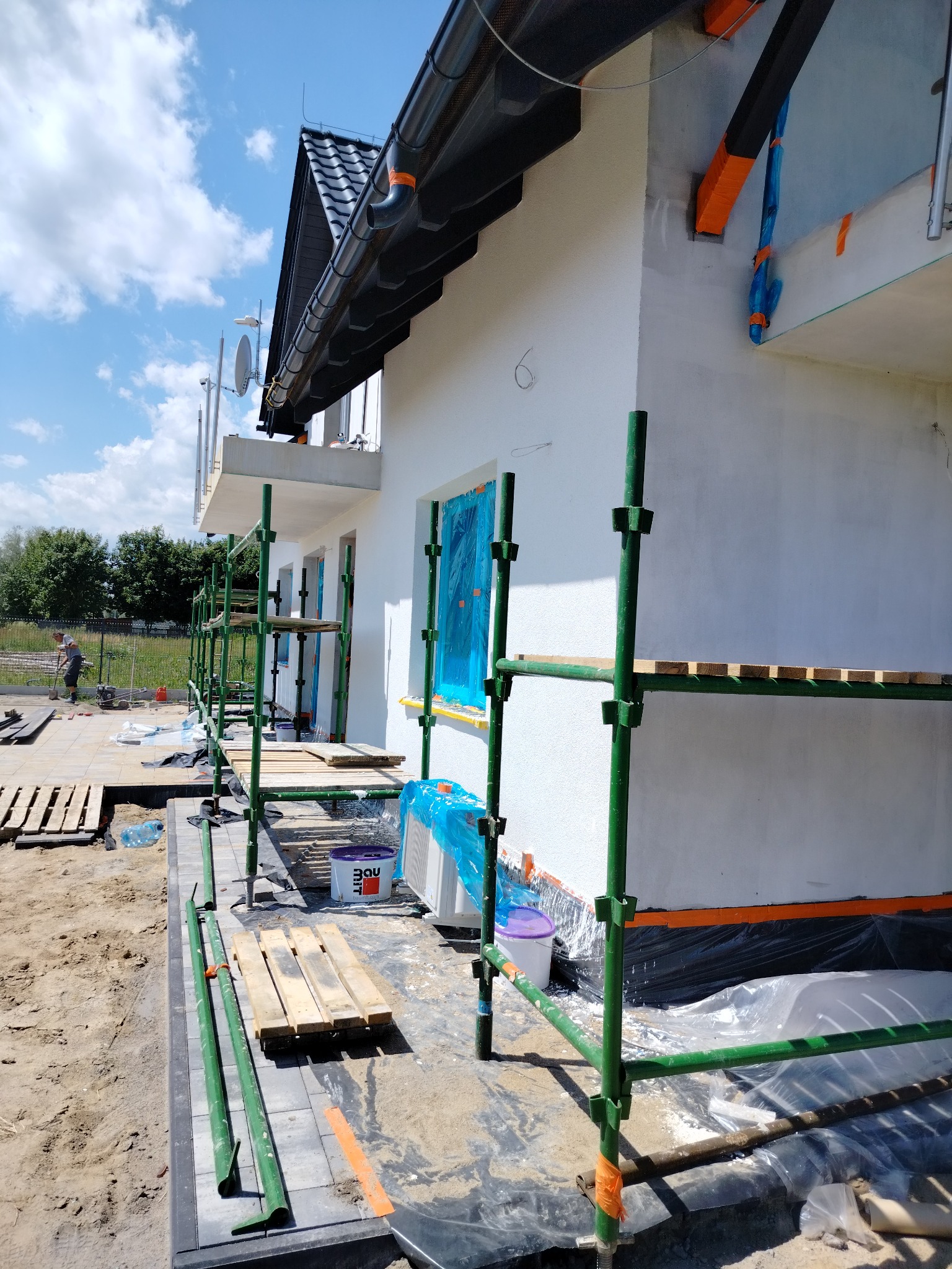 Dom jednorodzinny w trakcie prac elewacyjnych, widoczne rusztowania, zabezpieczone okna folią, ocieplenie fundamentów, pracownik w oddali.