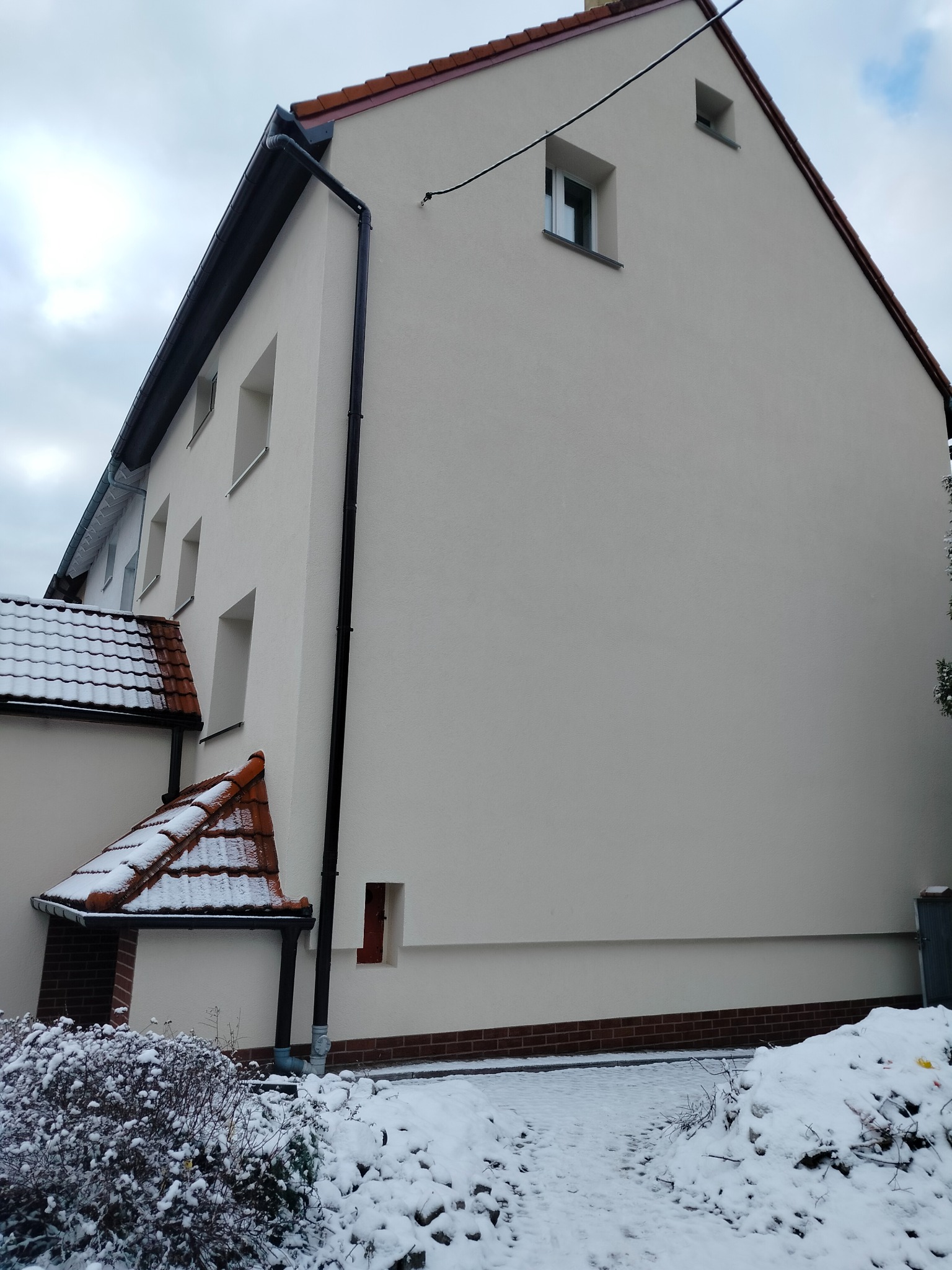 Świeżo otynkowana elewacja domu jednorodzinnego z widocznymi oknami i orynnowaniem w zimowej scenerii, z częściowo ośnieżonym dachem i ogrodem.