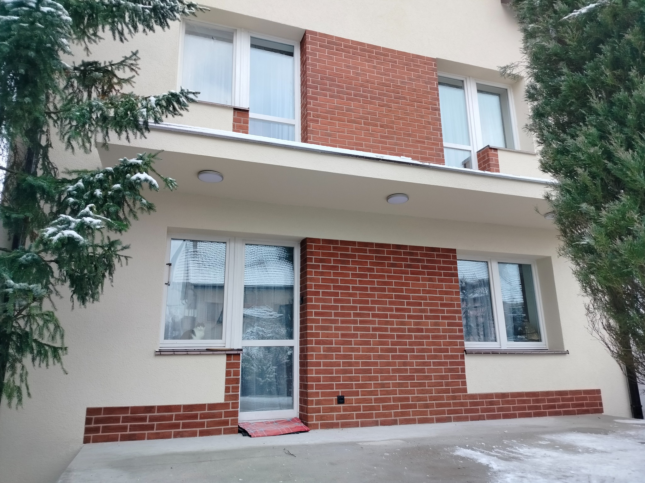 Dwupiętrowy dom z elewacją w kolorze kremowym i elementami z czerwonej cegły, widok z zewnątrz w zimowy dzień, z drzewami iglastymi po bokach i kotem w oknie.