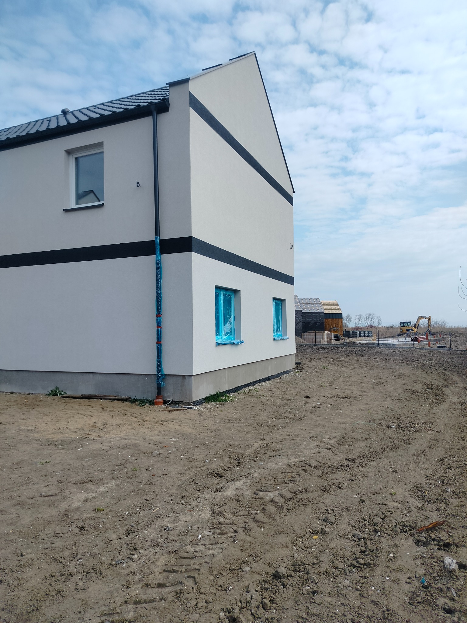 Nowoczesny dom jednorodzinny z jasną elewacją i czarnymi pasami dekoracyjnymi, okna zabezpieczone folią, widoczny teren budowy i maszyny w tle.