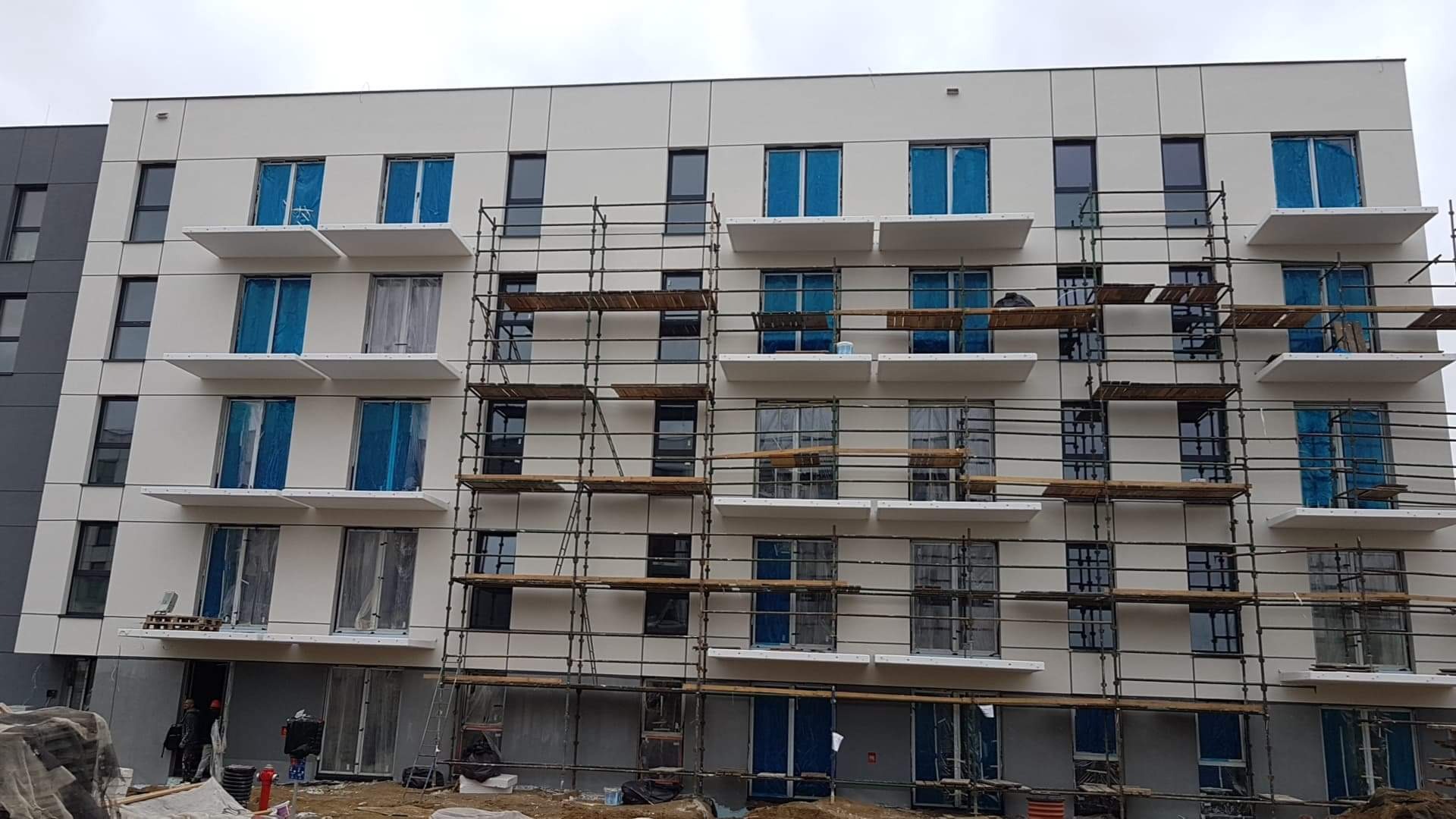 Wielopiętrowy budynek w trakcie prac elewacyjnych, z rusztowaniami i oknami zabezpieczonymi folią, widoczne balkony.