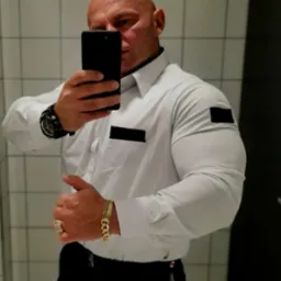 Mężczyzna w białej koszuli z czarnymi naszywkami robi selfie w łazience z białymi kafelkami, widoczny złoty zegarek i bransoleta.