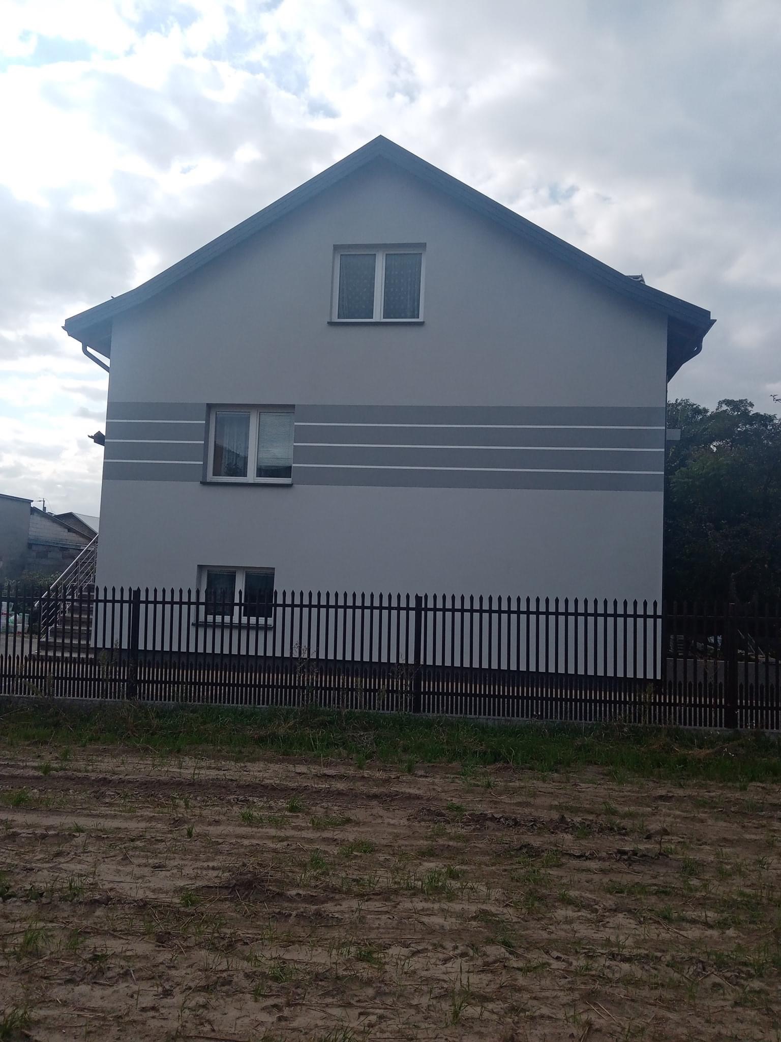 Dwukondygnacyjny dom z szarą elewacją i poziomymi pasami, widoczny z poziomu gruntu, ogrodzony czarnym metalowym płotem na tle pochmurnego nieba.