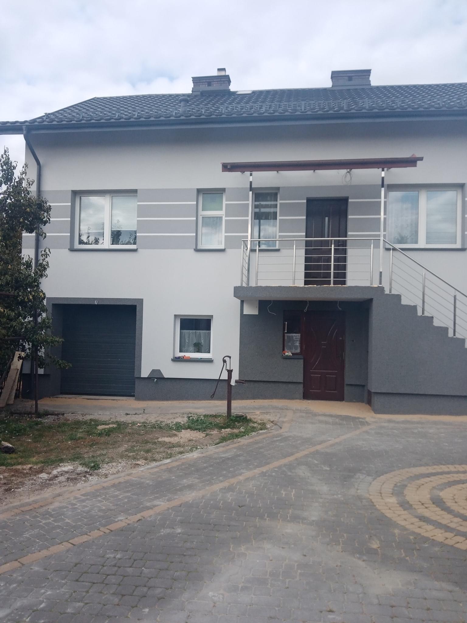 Dwukondygnacyjny dom z szarą elewacją, widoczne wejście na piętro po schodach zewnętrznych z metalową barierką, garaż z bramą segmentową i chodnik z kostki brukowej.