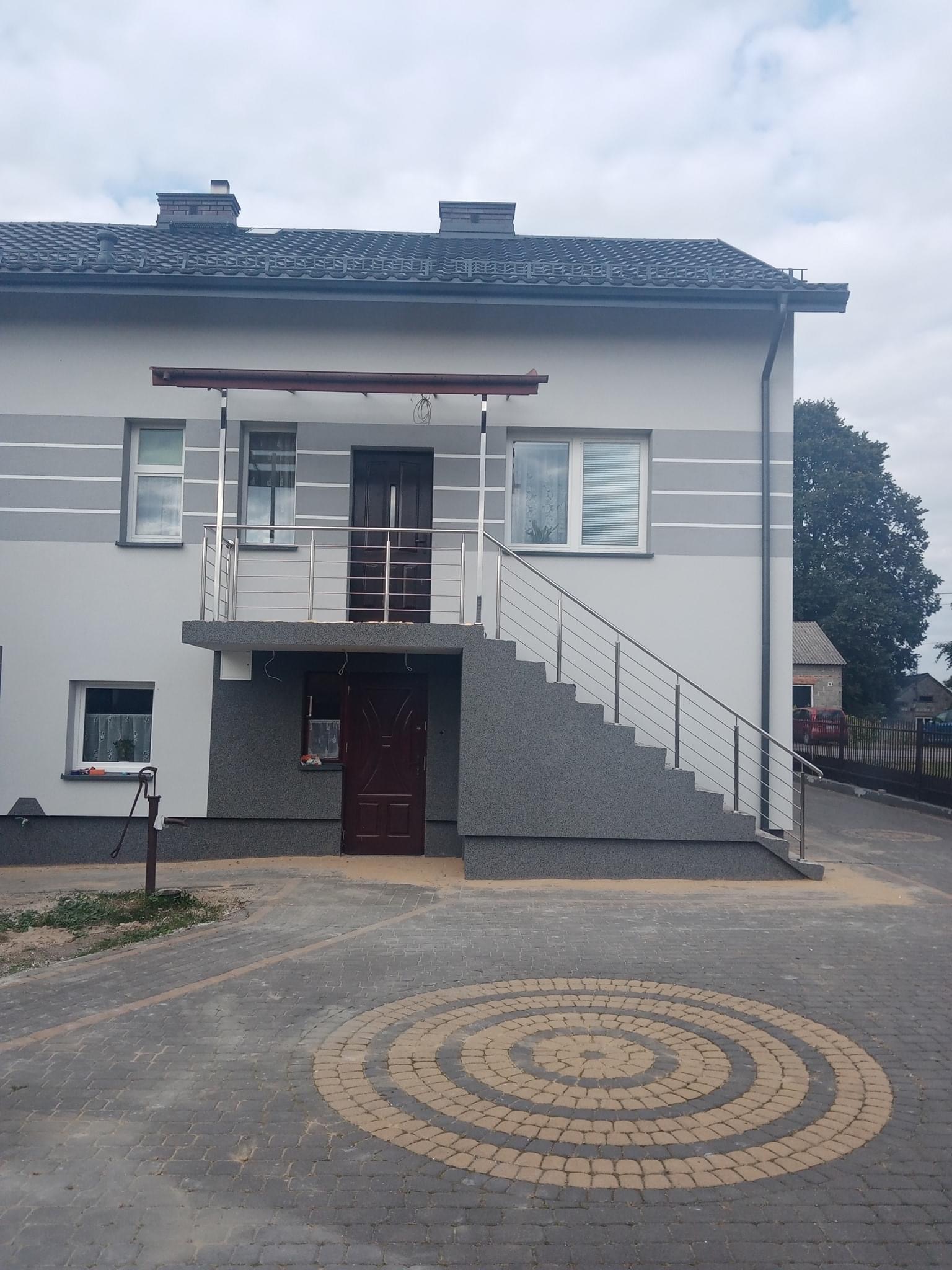 Dwukondygnacyjny dom z szarą elewacją i poziomymi pasami, schody zewnętrzne z metalową balustradą, dekoracyjny bruk z okręgami na podjeździe.