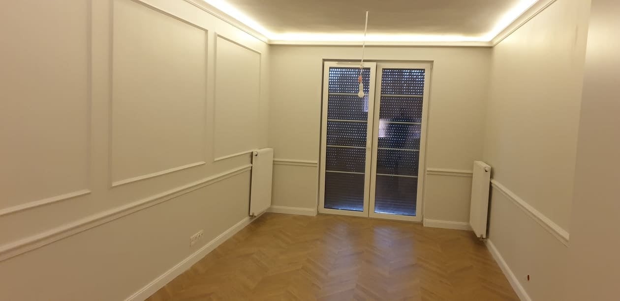 Jasne wnętrze po remoncie z dekoracyjnymi listwami ściennymi, parkietem w jodełkę i oświetleniem LED podsufitowym.