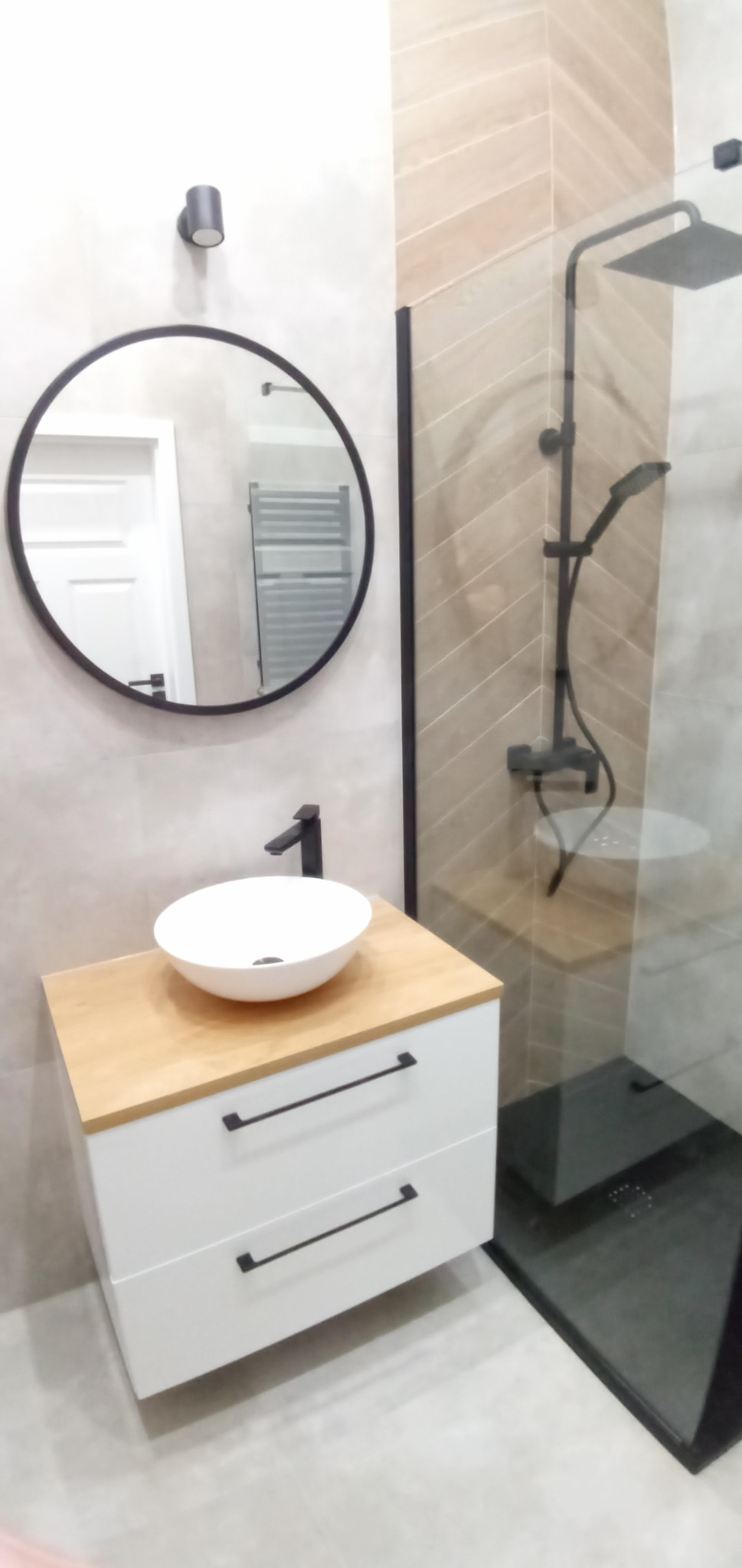 Nowoczesna łazienka z okrągłym lustrem, białą szafką pod umywalkę z drewnianym blatem, czarną armaturą i kabiną prysznicową typu walk-in z czarnymi elementami.