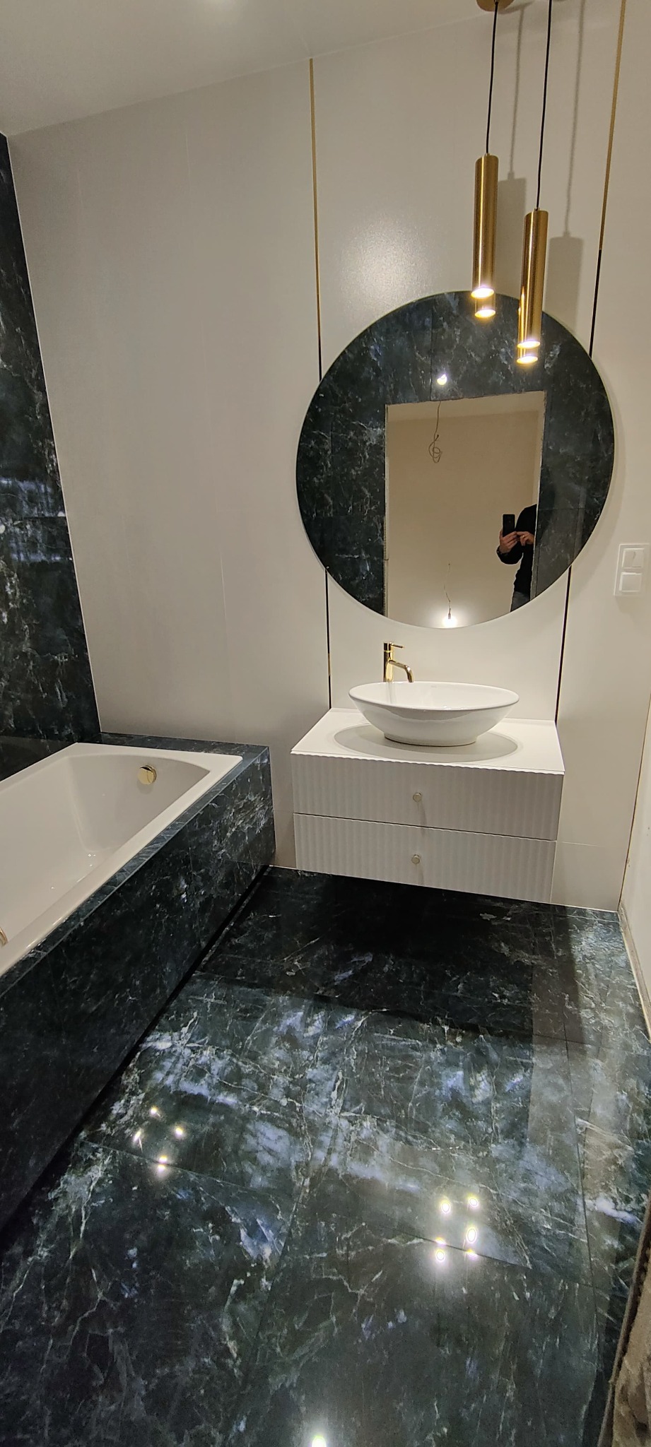 Nowoczesna łazienka z ciemnozielonymi marmurowymi płytkami na podłodze i ścianie, owalne lustro z ramą z tego samego materiału, biała umywalka nablatowa z złotą baterią i biała szafka pod umywalką...