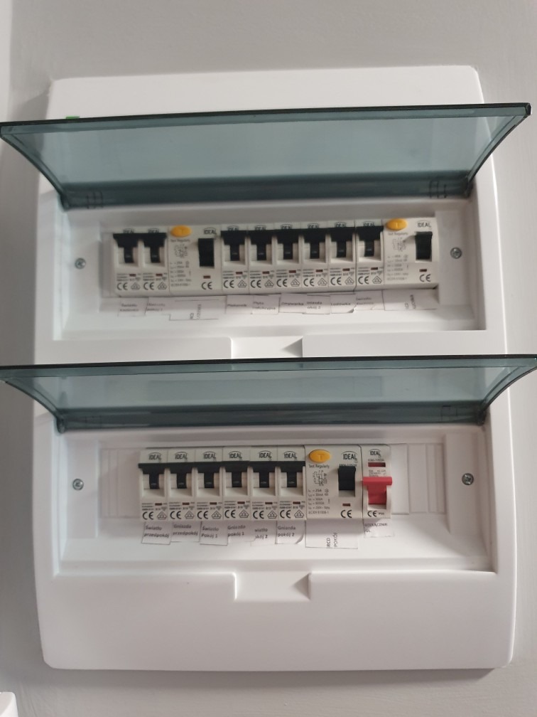 Rozdzielnica elektryczna z dwoma rzędami bezpieczników marki Ideal, z uchylnymi, przezroczystymi osłonami, zamontowana na białej ścianie. Jeden wyłącznik w pozycji wyłączonej.