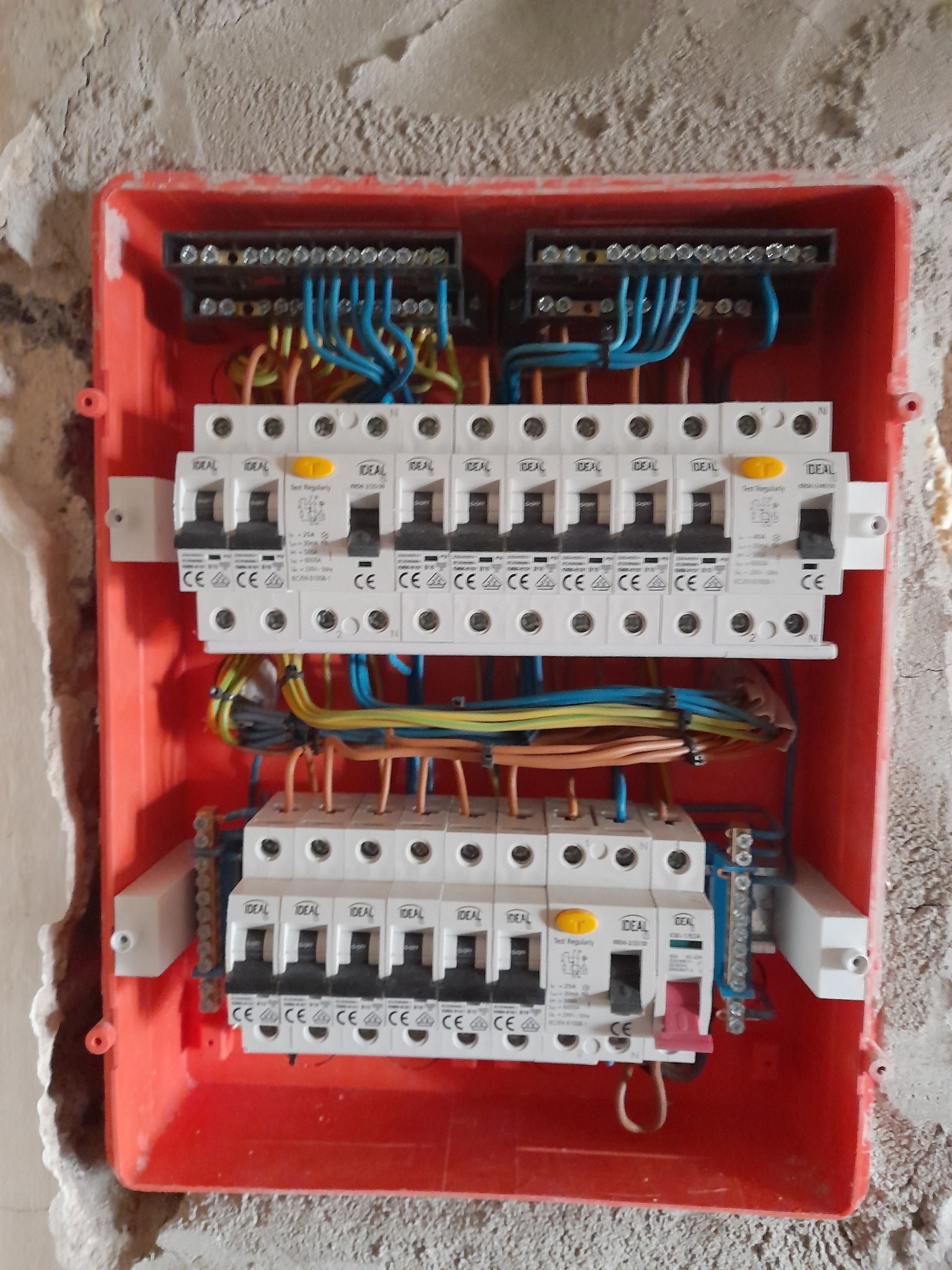 Otwarta, czerwona skrzynka rozdzielcza z zamontowanymi bezpiecznikami i podłączonymi przewodami w surowym otoczeniu ściany z widocznymi śladami tynku.