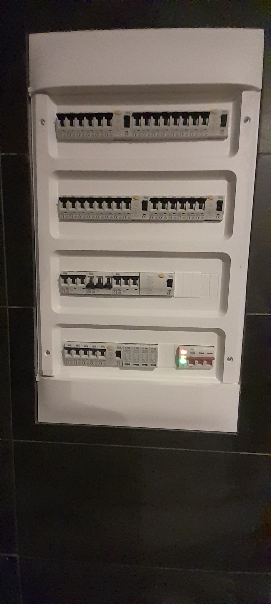 Biała skrzynka rozdzielcza z bezpiecznikami i wyłącznikami różnicowoprądowymi zamontowana na ciemnej, prawdopodobnie kafelkowanej ścianie, z widoczną diodą LED sygnalizującą stan pracy jednego...