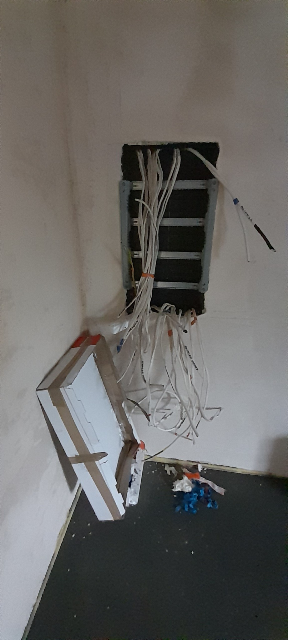 Wystające ze ściany przewody elektryczne, częściowo zabezpieczone taśmą, widoczne elementy montażowe i resztki materiałów budowlanych na podłodze.