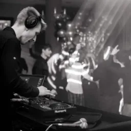 Czarno-białe zdjęcie DJ-a w słuchawkach miksującego muzykę na konsoli Numark, w tle rozmazane sylwetki bawiących się ludzi i światła sceniczne.