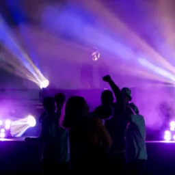 DJ za konsoletą grający muzykę dla tańczącego tłumu w fioletowym oświetleniu scenicznym i sztucznym dymie.