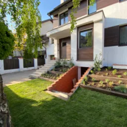 Nowoczesny dom z białymi ścianami i brązowymi panelami, otoczony świeżo założonym trawnikiem, niskimi rabatami z nowymi nasadzeniami i ceglanym wejściem do piwnicy.