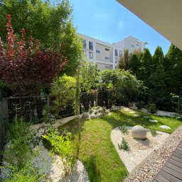 INTELLIGARDEN ALICJA KAMIŃSKA - Mały, nowoczesny ogród z trawnikiem, białymi kamykami, ścieżką z kamiennych płyt i dekoracyjną kulą, otoczony zielenią i budynkami mieszkalnymi.