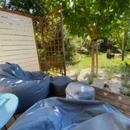 Relaksująca strefa wypoczynkowa na drewnianym tarasie z szarymi pufami typu sako, białym ażurowym panelem i drewnianą kratownicą, otoczona zielenią trawnika i drzew.