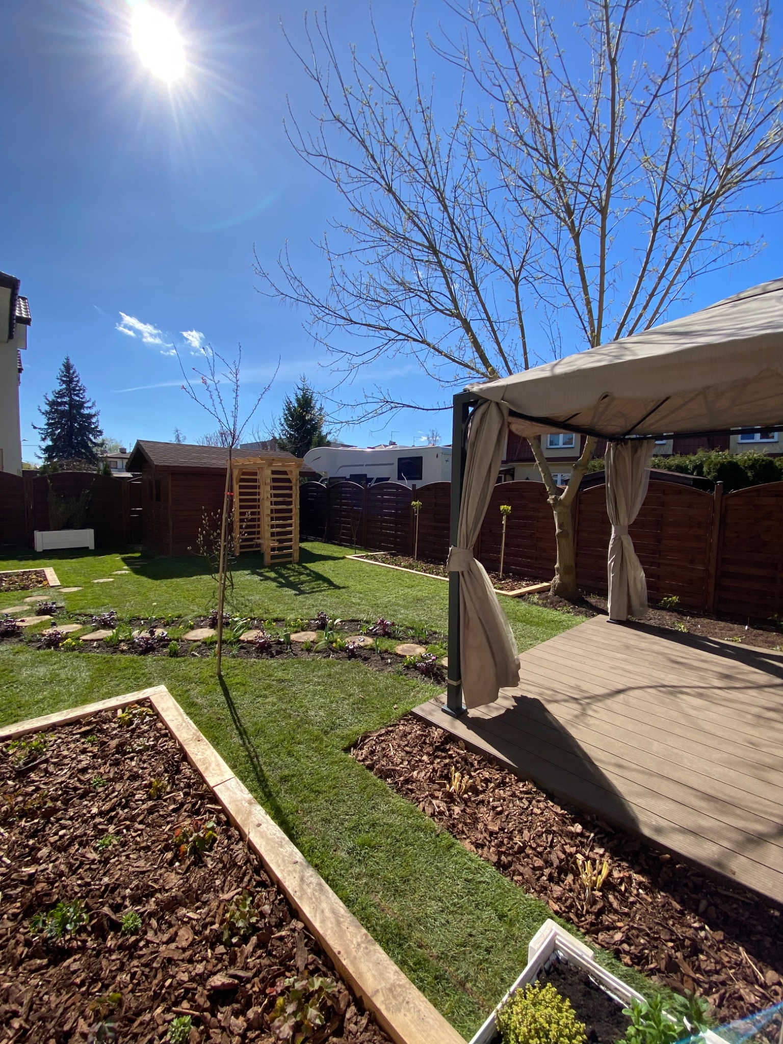 Ujęcie na ogród z drewnianą altaną z zasłonami, drewnianym domkiem narzędziowym i podwyższonymi rabatami z korą, świeżo założony trawnik, słoneczny dzień.