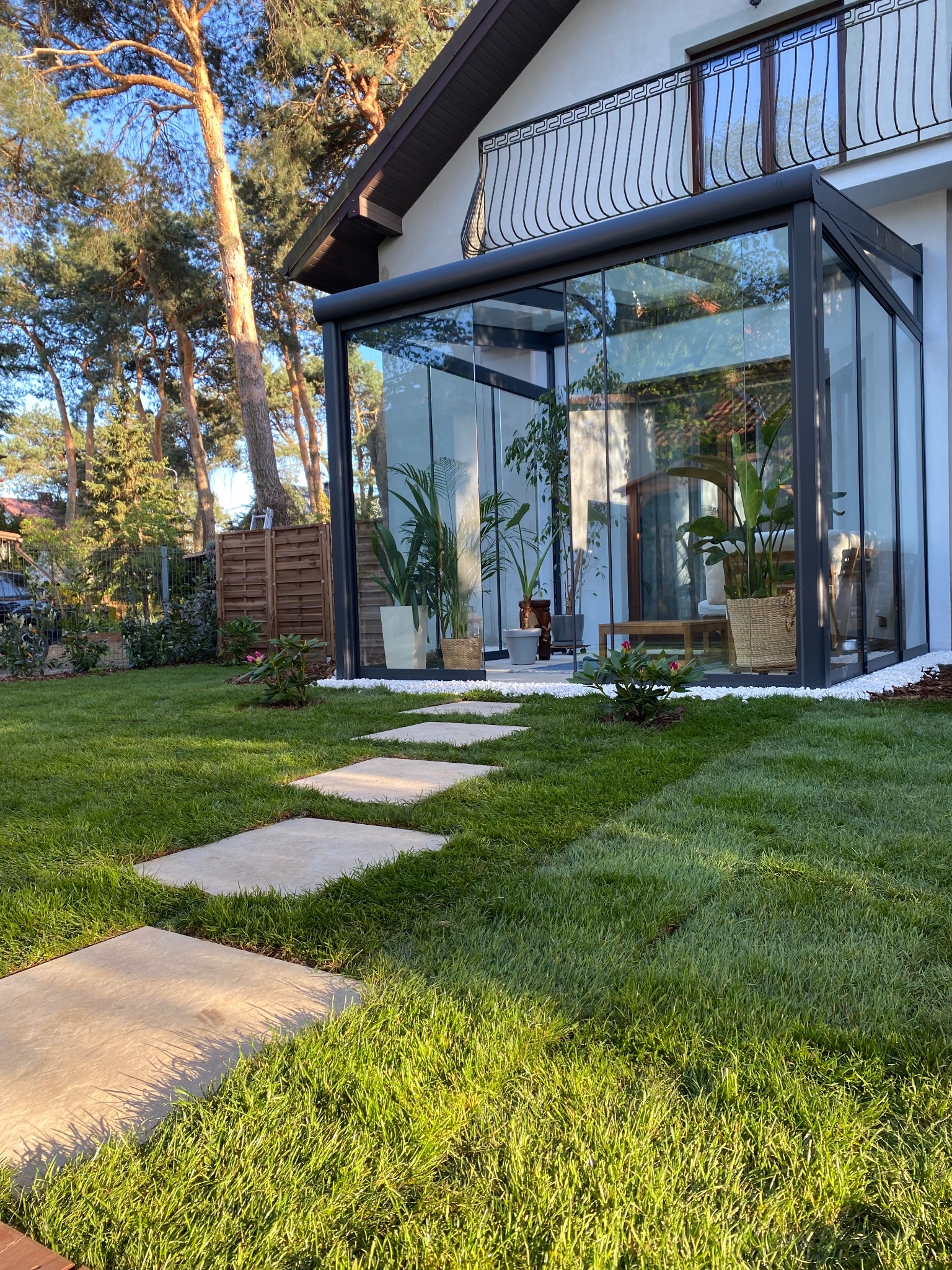 Nowoczesny ogród z równo przystrzyżonym trawnikiem, ścieżką z płyt betonowych prowadzącą do przeszklonej oranżerii z roślinami doniczkowymi, otoczonej białym kamieniem, w tle drzewa iglaste.