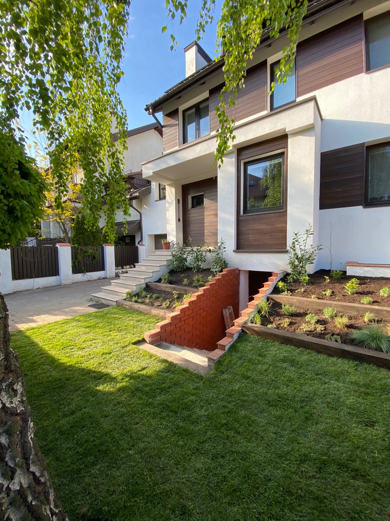 Nowoczesny dom z białymi ścianami i brązowymi panelami, otoczony świeżo założonym trawnikiem, niskimi rabatami z nowymi nasadzeniami i ceglanym wejściem do piwnicy.