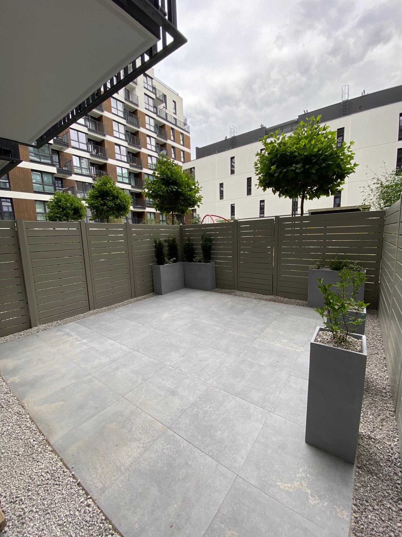 Nowoczesny taras wyłożony szarą płytką z minimalistycznym ogrodzeniem z poziomych desek i donicami z zielenią na tle miejskiej zabudowy.