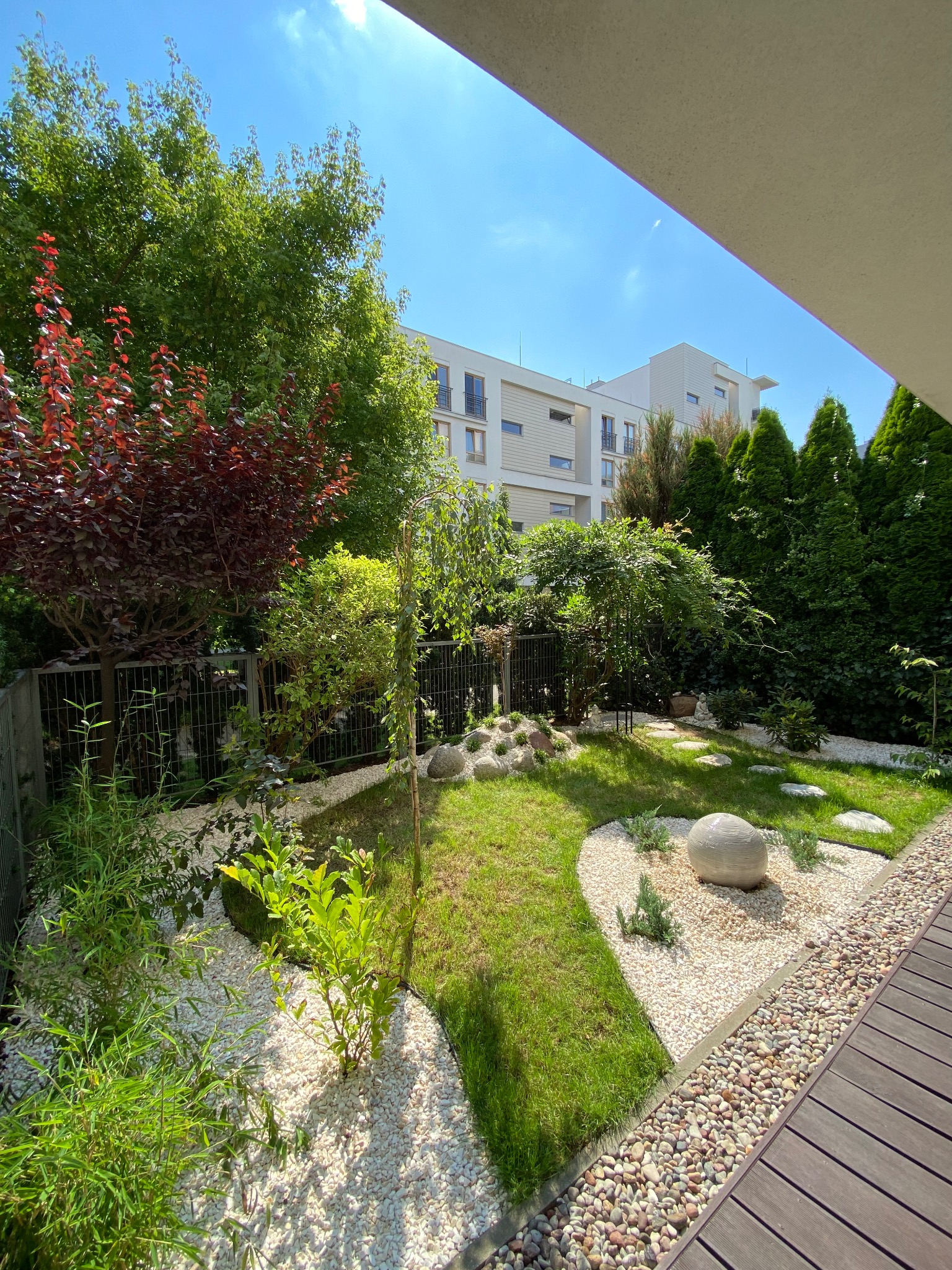 Mały, nowoczesny ogród z trawnikiem, białymi kamykami, ścieżką z kamiennych płyt i dekoracyjną kulą, otoczony zielenią i budynkami mieszkalnymi.