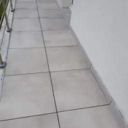 Jasnoszare płytki gresowe ułożone na balkonie, widoczne fugi, balustrada metalowa i fragment elewacji budynku w tle.