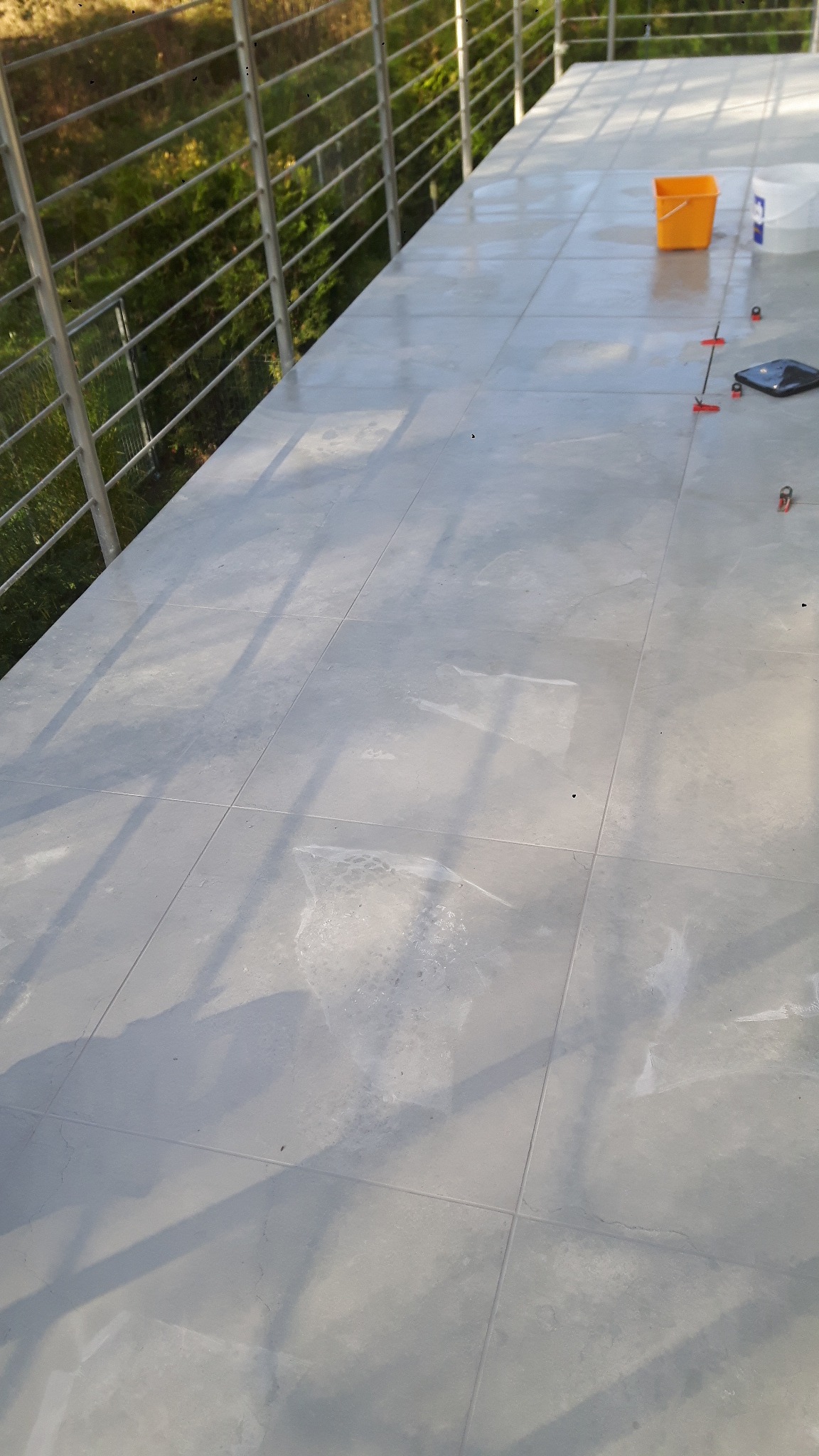 Świeżo ułożone, jasnoszare płytki na balkonie, widoczne narzędzia glazurnicze, wiadro i system poziomowania płytek.