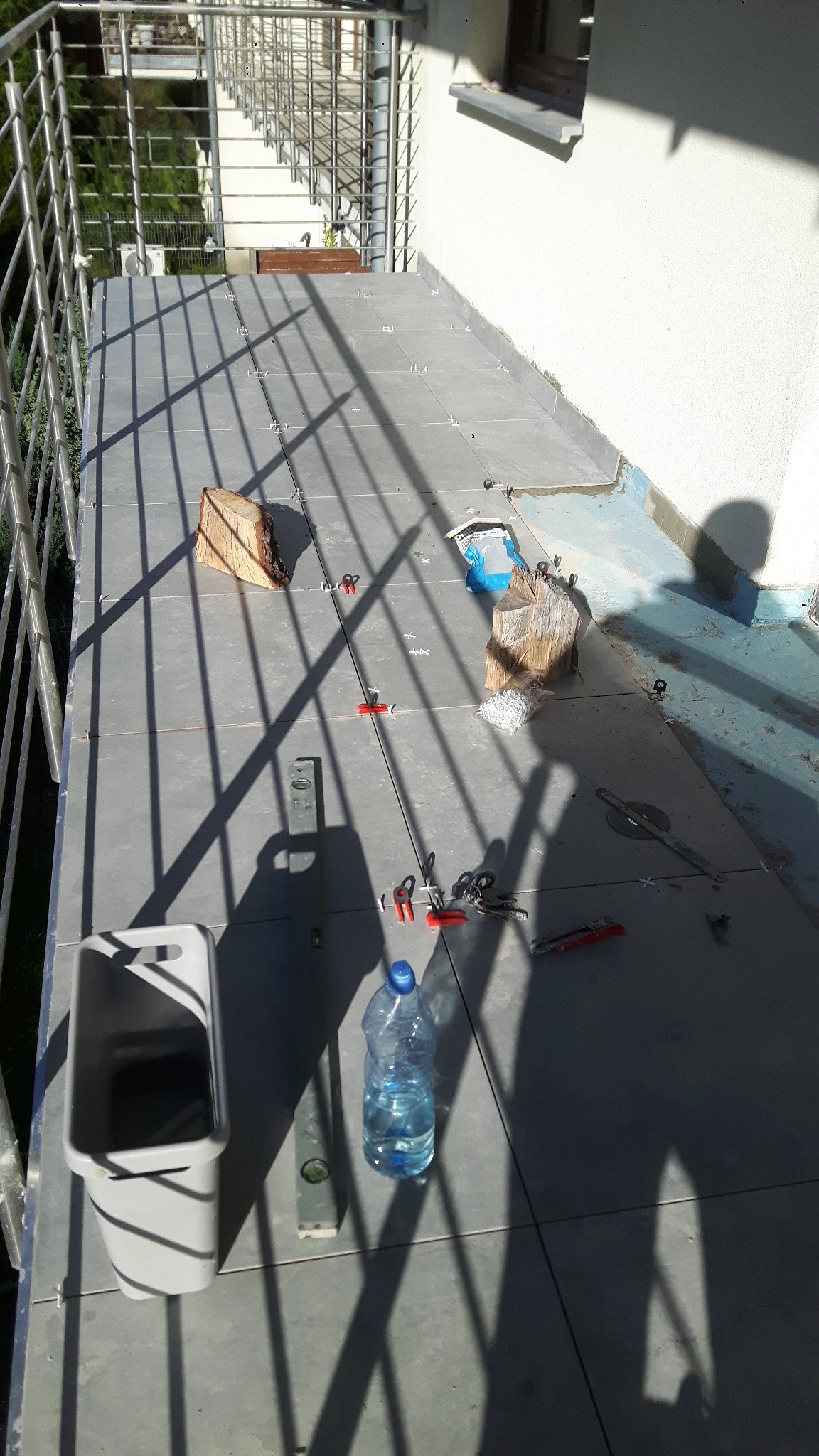 Szare płytki na balkonie w trakcie układania, widoczne poziomica, butelka wody, wiadro i kliny do płytek, cień barierki