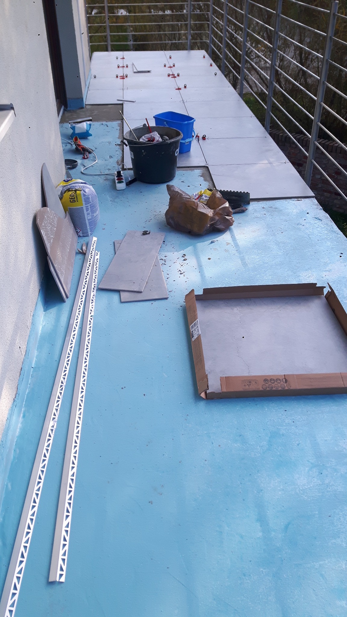 Balkon w trakcie układania płytek, widoczne płytki, poziomice, wiadro z zaprawą, klej, listwy wykończeniowe i otwarte opakowanie płytek.