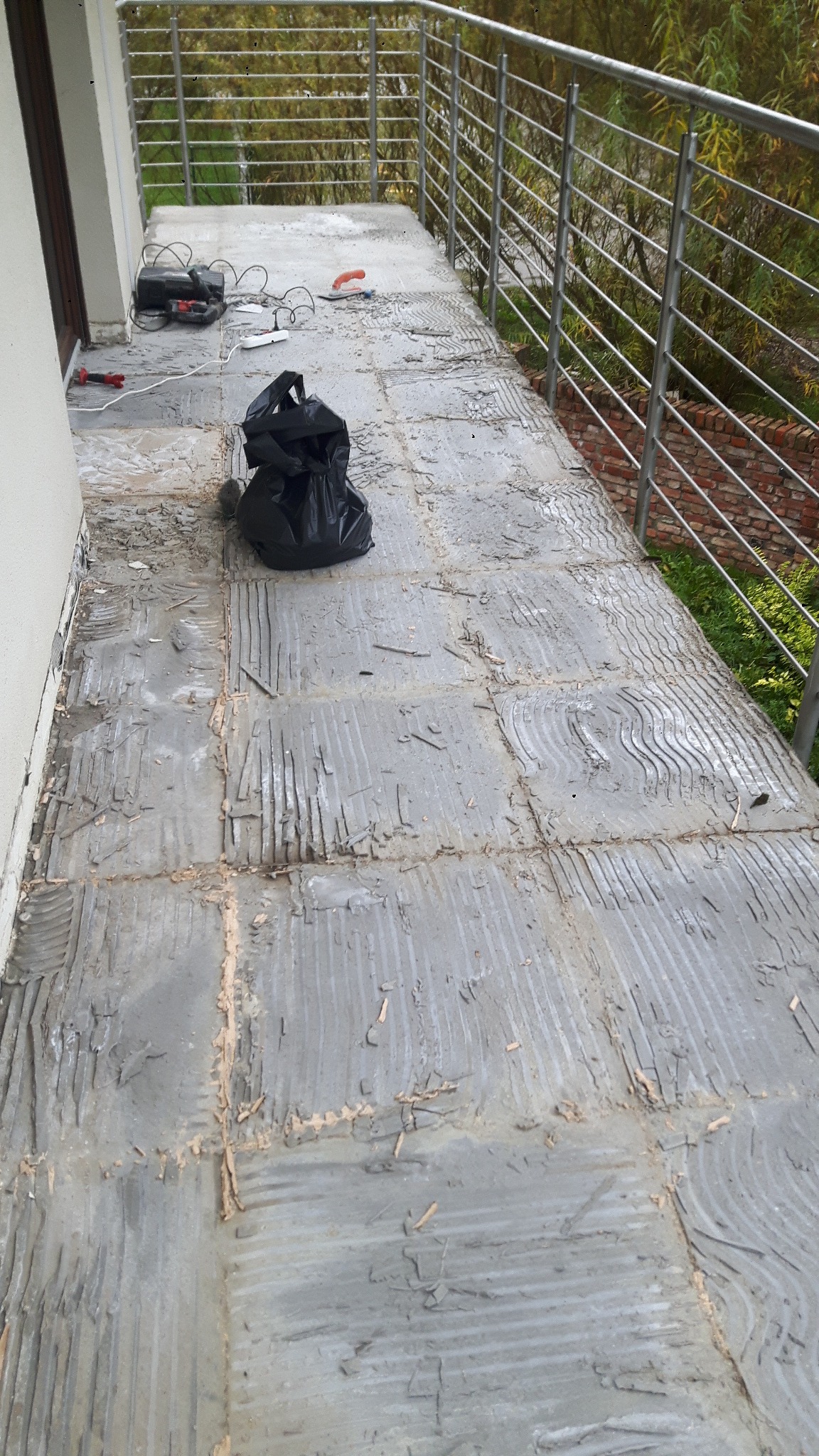 Demontaż płytek na balkonie, widoczne pozostałości kleju, narzędzia i worek na gruz, balustrada ze stali nierdzewnej w tle.