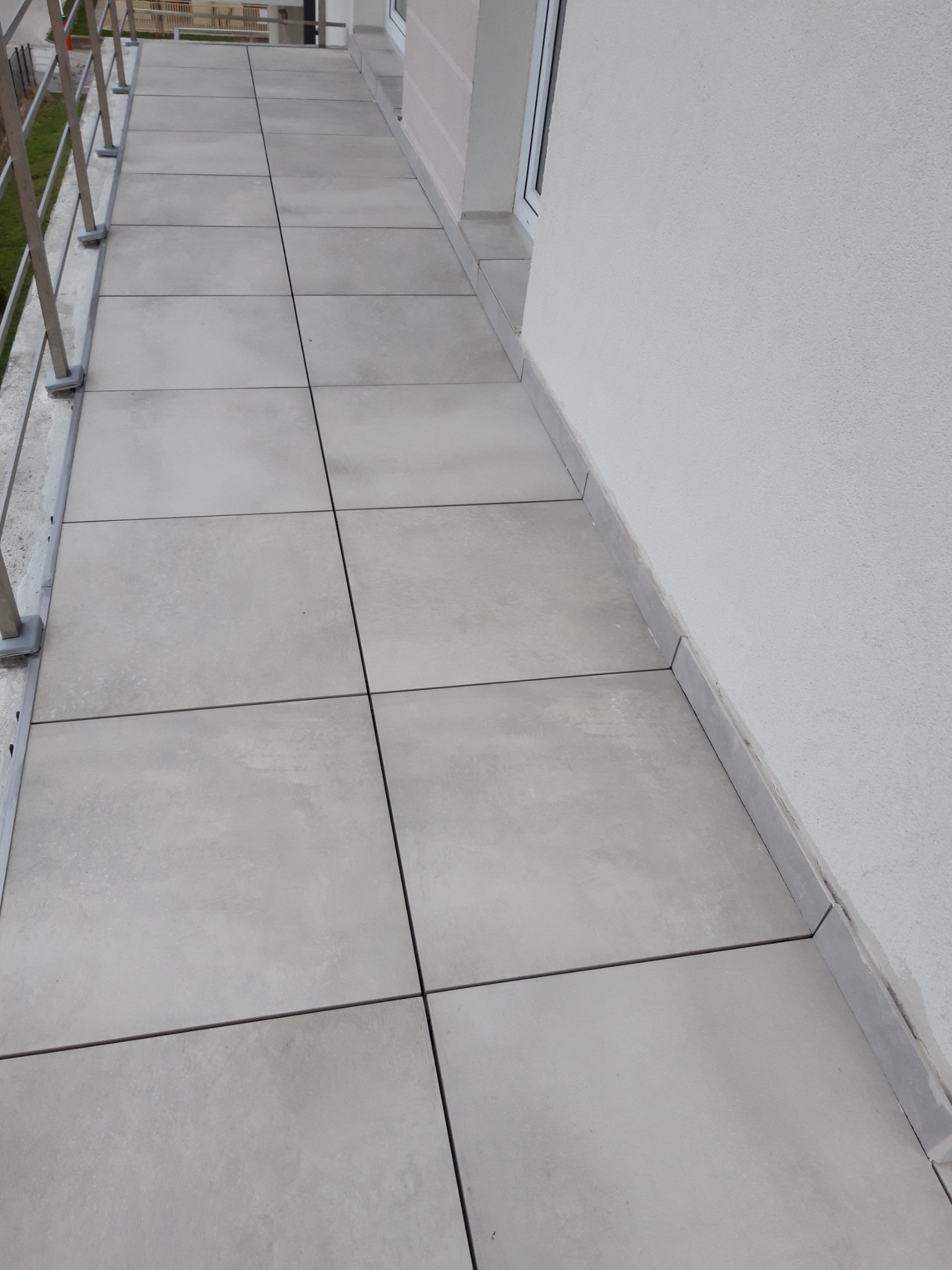 Jasnoszare płytki gresowe ułożone na balkonie, widoczne fugi, balustrada metalowa i fragment elewacji budynku w tle.