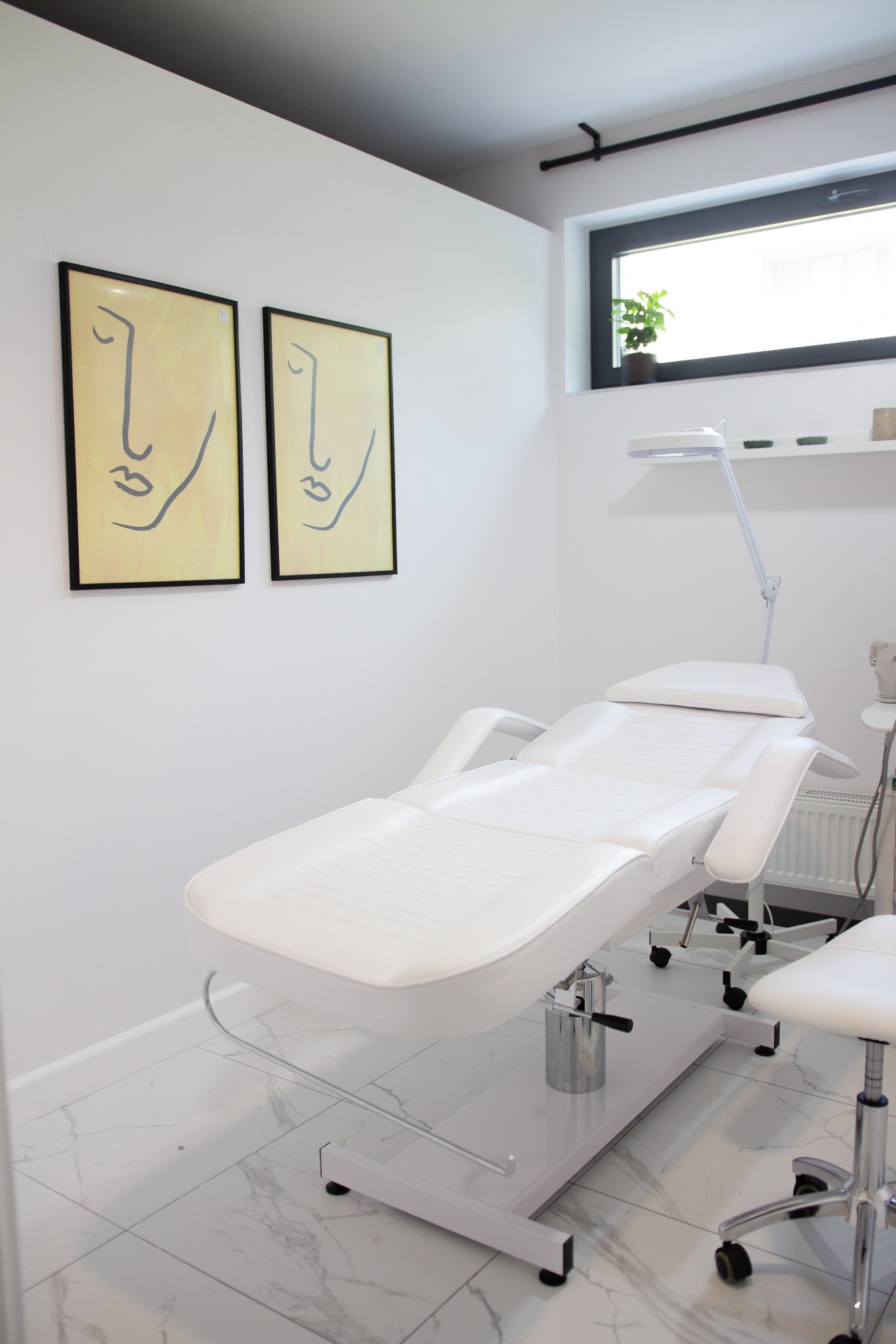 Jasne wnętrze gabinetu kosmetycznego z białym, regulowanym fotelem do zabiegów, lampą kosmetyczną i minimalistycznymi obrazami twarzy na ścianie.