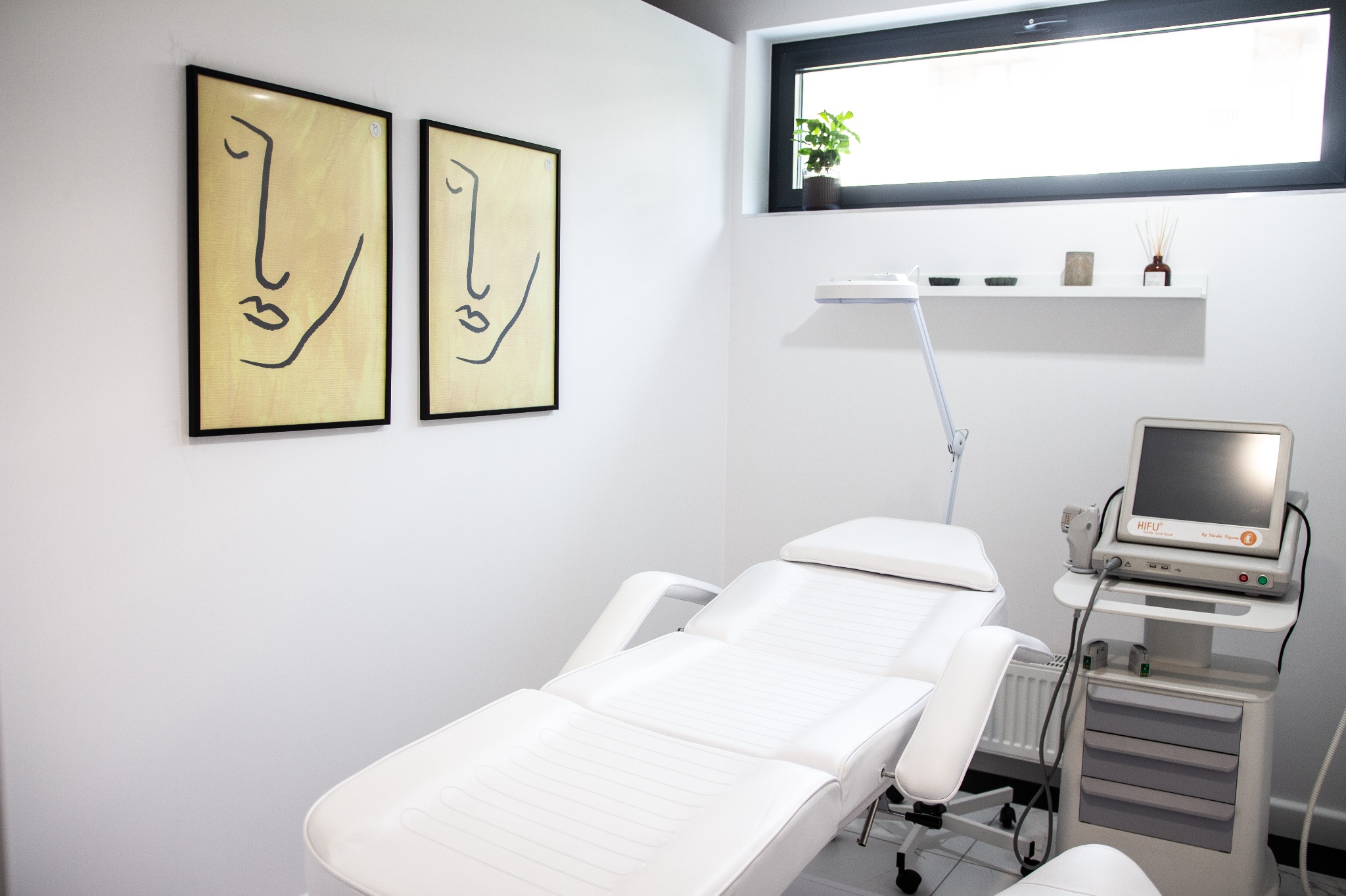 Biały fotel kosmetyczny z regulowanym oparciem i podłokietnikami, obok urządzenie HIFU na kółkach, minimalistyczne obrazy na ścianie, roślina w doniczce na parapecie okna