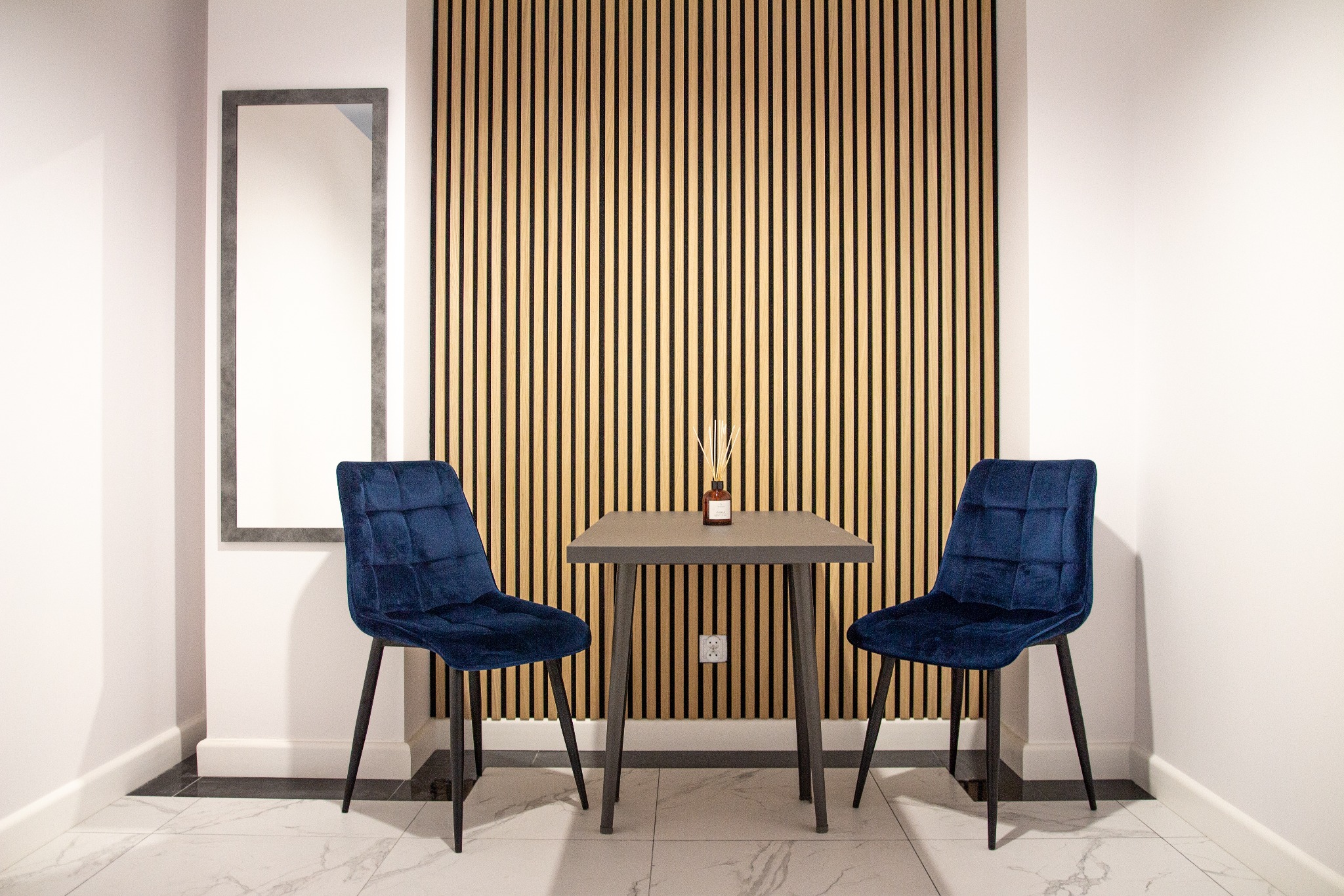 Nowoczesne wykończenie wnętrza lokalu usługowego: ciemnoniebieskie, welurowe krzesła przy szarym stoliku z dyfuzorem zapachowym na tle ściany z drewnianych paneli i lustra.