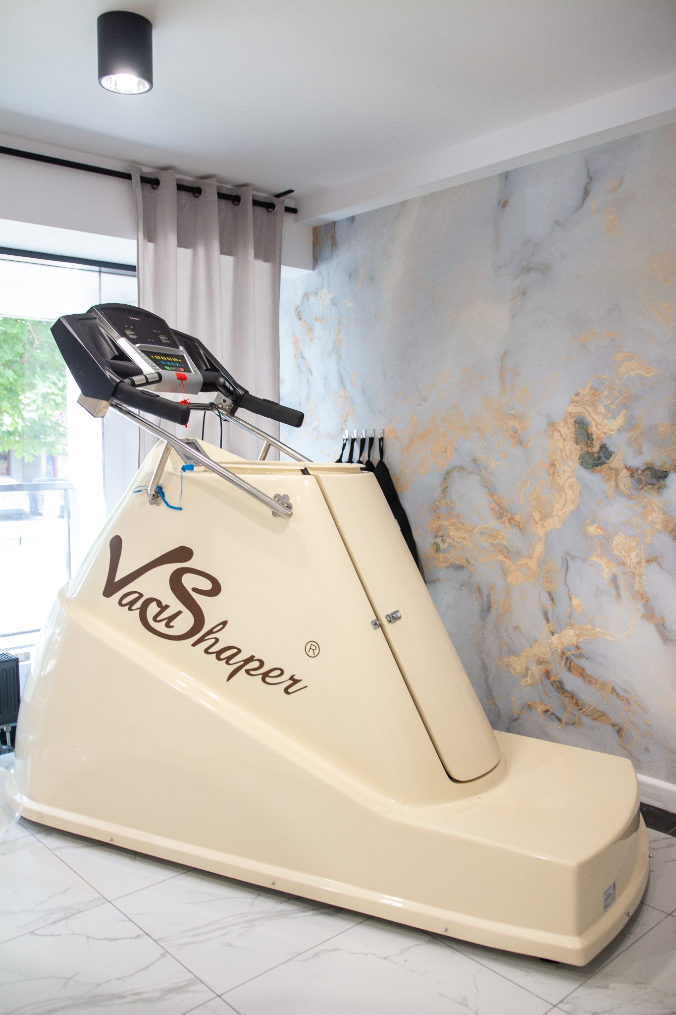 Vacu Shaper, urządzenie do ćwiczeń z podciśnieniem, w jasnym pomieszczeniu z marmurową ścianą i szarymi zasłonami.