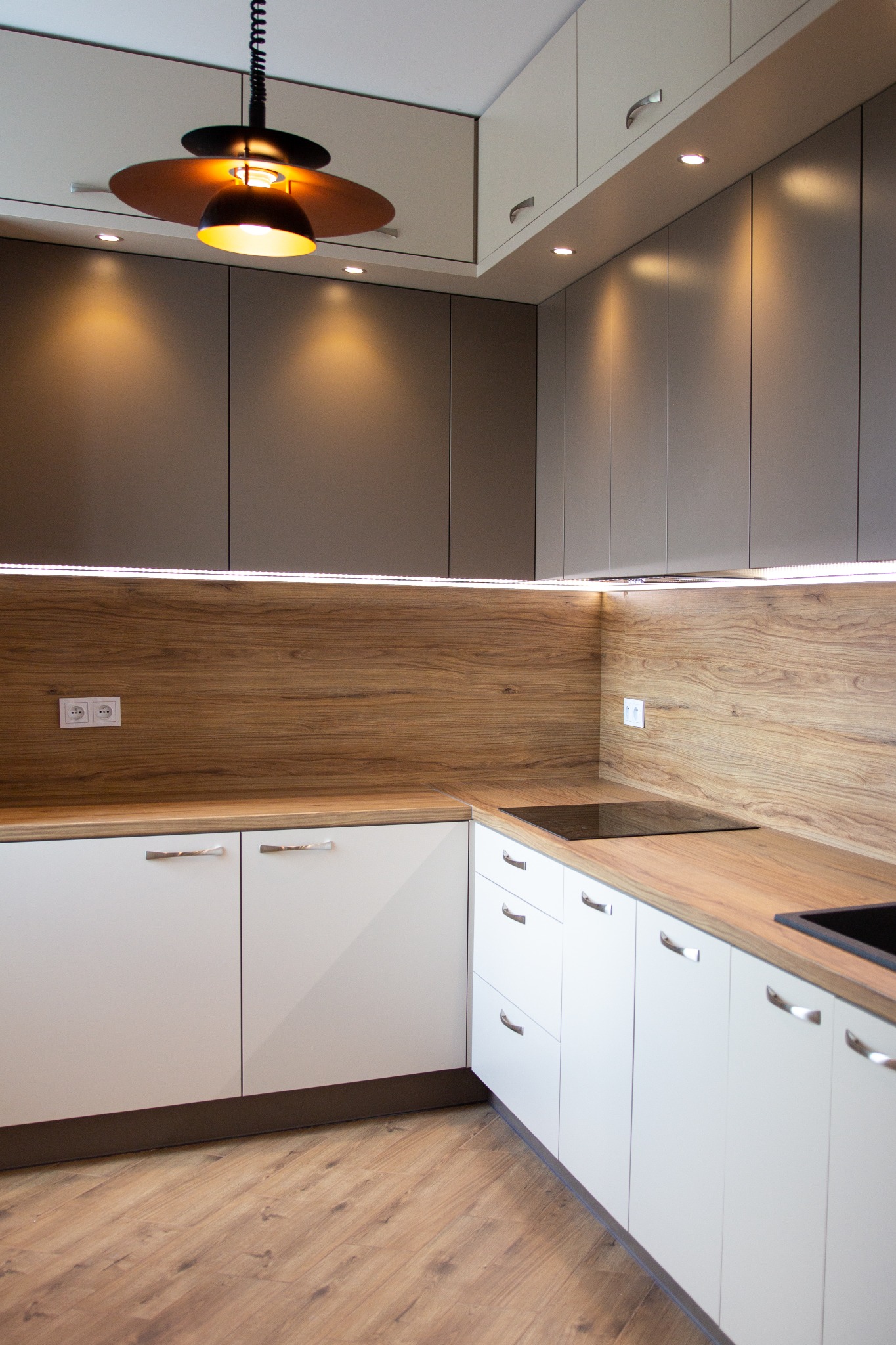Nowoczesna kuchnia z białymi szafkami dolnymi, szarymi górnymi, drewnianym blatem i panelem ściennym, oświetlona listwą LED i wiszącą lampą z brązowym kloszem.