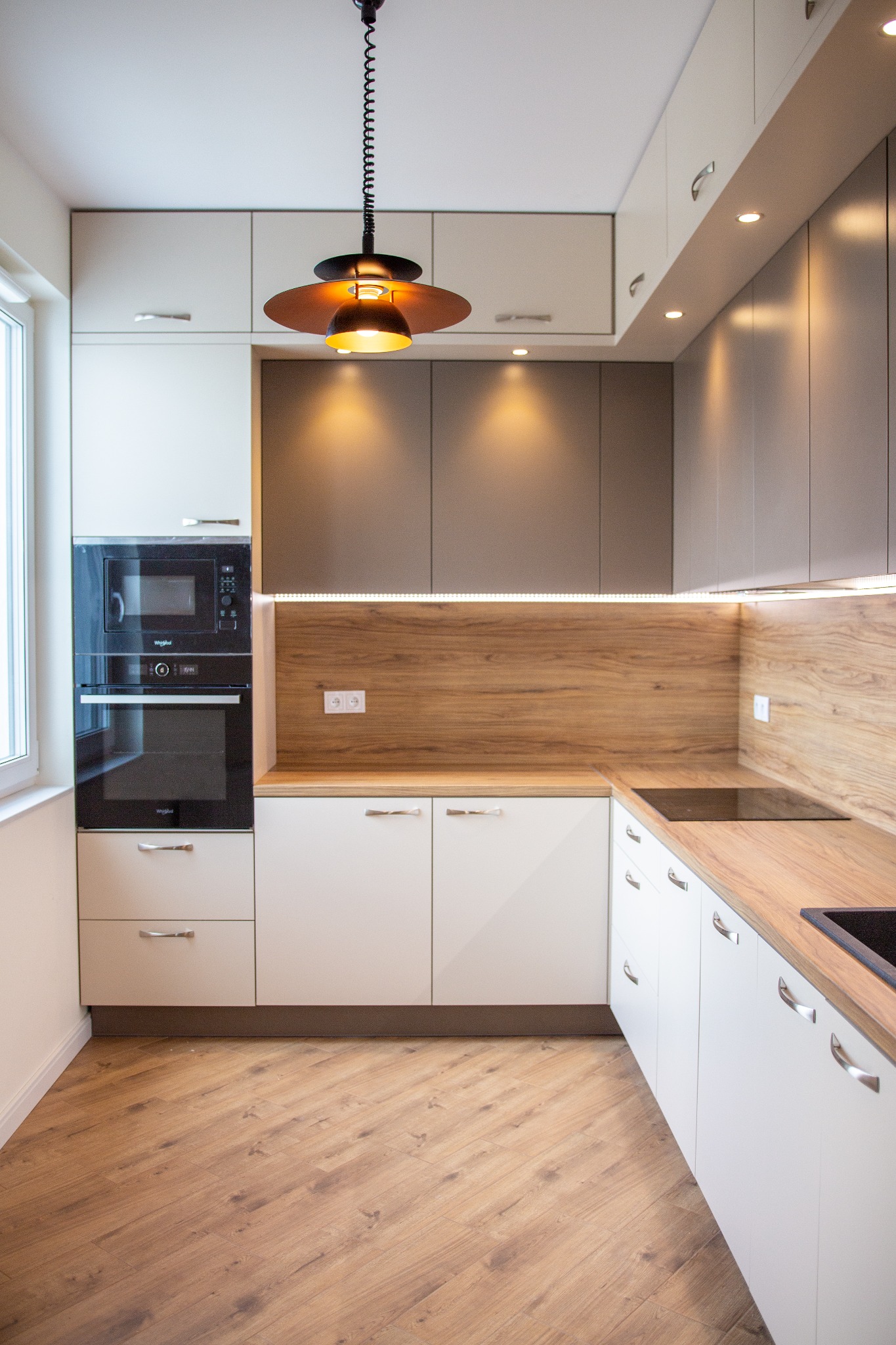 Nowoczesna kuchnia z białymi szafkami, drewnianym blatem i brązowymi akcentami, oświetlona lampą wiszącą w stylu retro i ledami pod szafkami górnymi.
