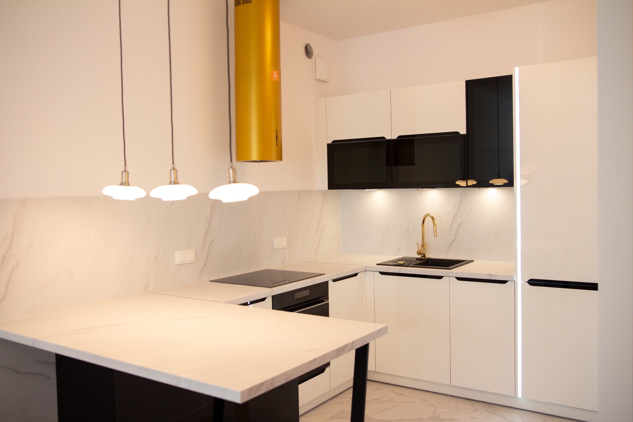 Nowoczesna, biała kuchnia z czarnymi akcentami, złotą armaturą i okapem. Widoczne trzy lampy wiszące nad blatem. Marmurowy wzór na ścianie i podłodze.