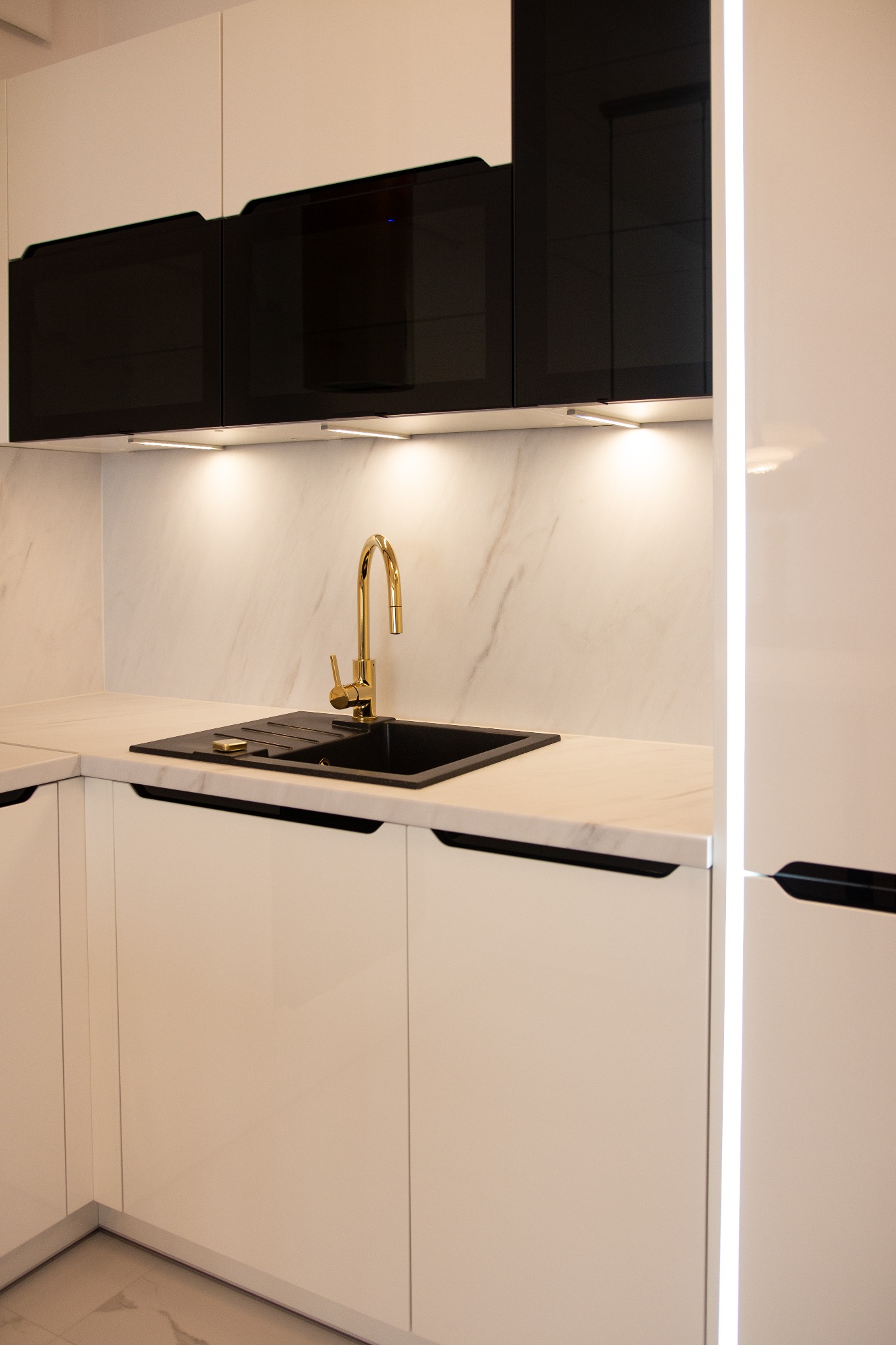 Nowoczesna kuchnia: białe szafki z czarnymi akcentami, złota bateria przy czarnym zlewozmywaku, marmurowy backsplash, oświetlenie LED pod szafkami wiszącymi.