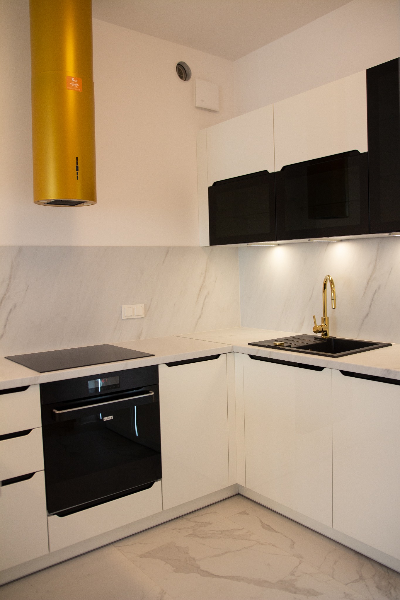 Nowoczesna kuchnia z białymi szafkami, czarnymi akcentami, złotą armaturą i okapem w kolorze złota.