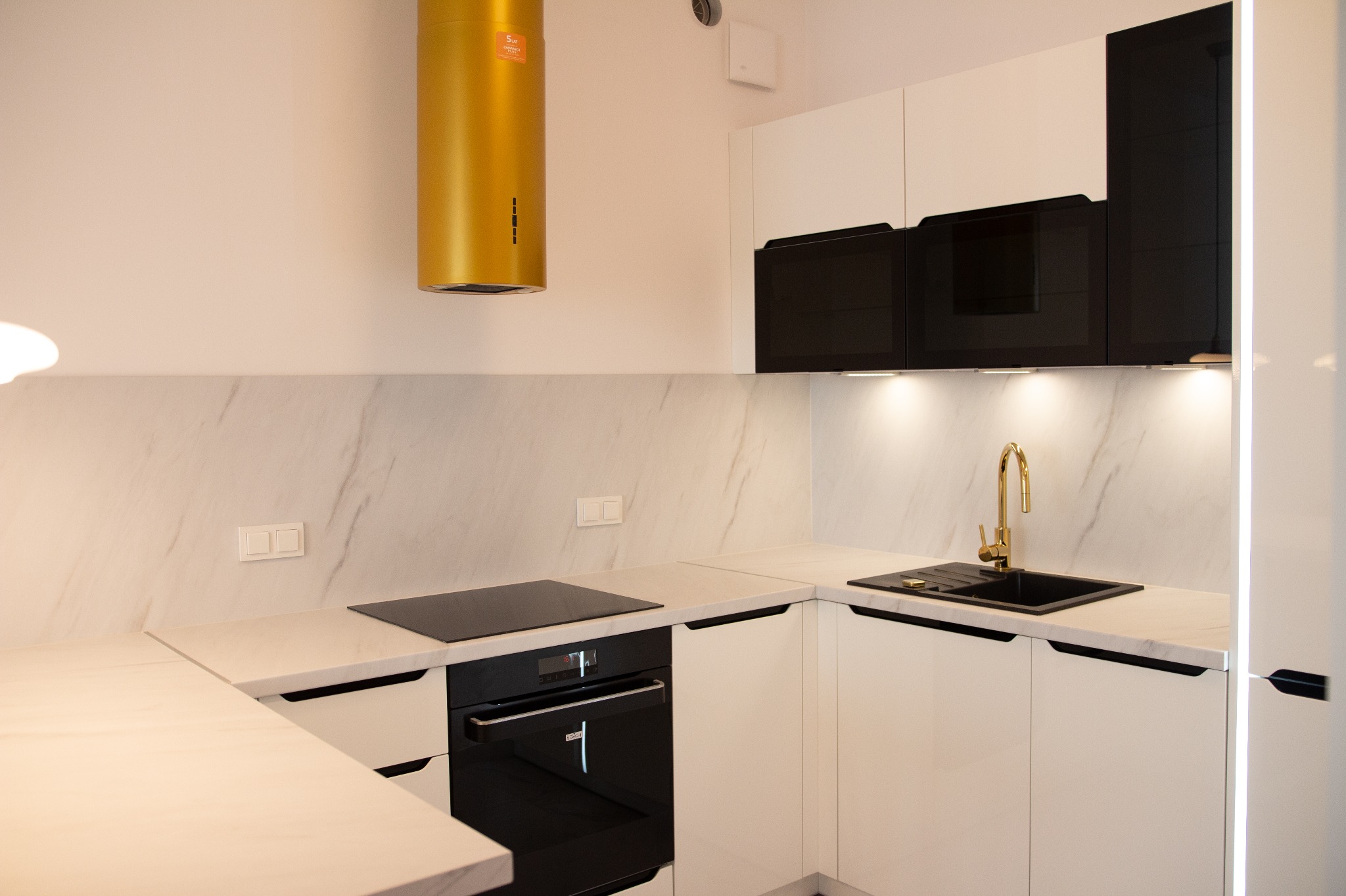Nowoczesna kuchnia z białymi szafkami, czarnym sprzętem AGD i złotymi akcentami: bateria i okap.