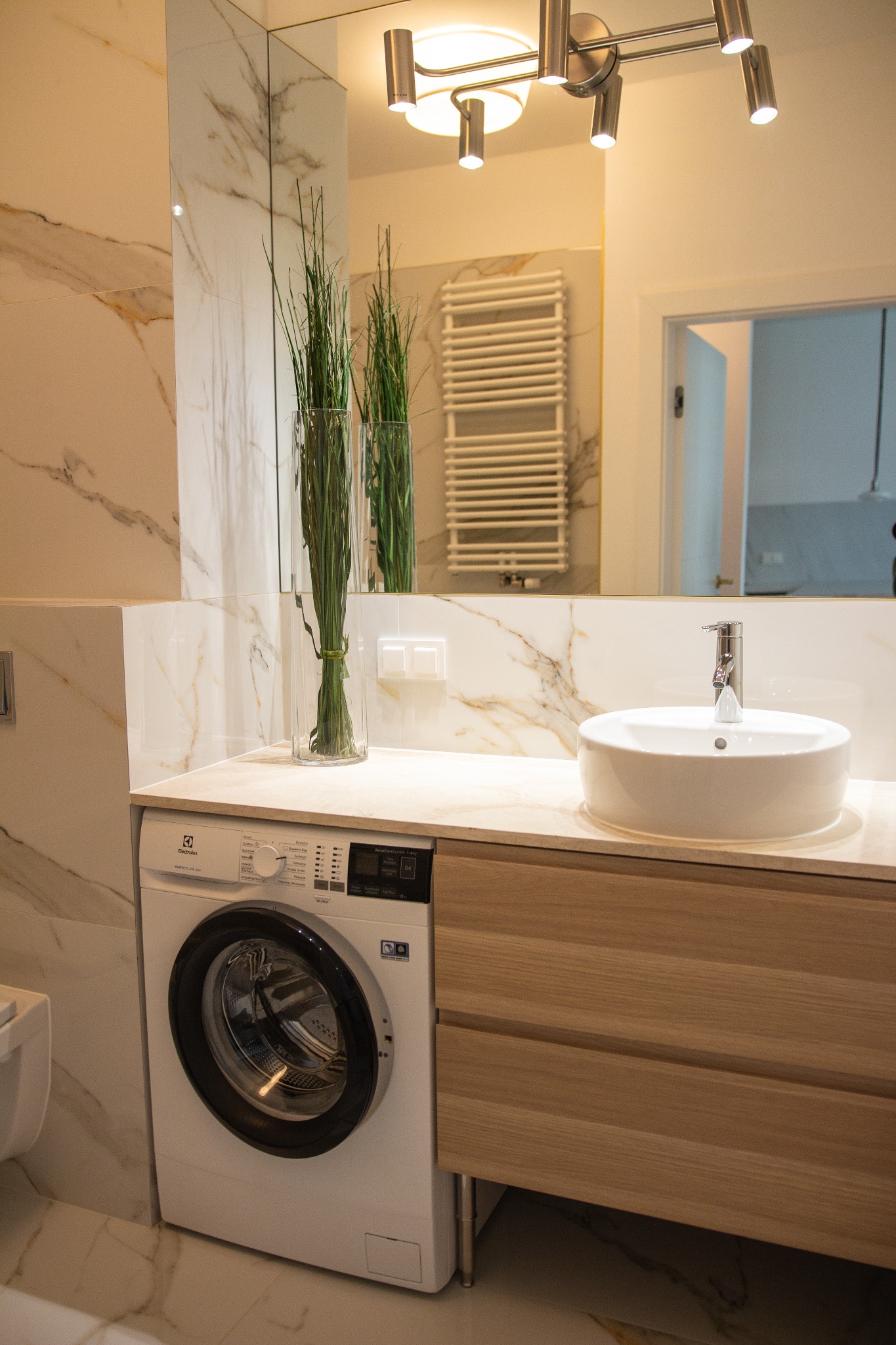 Nowoczesna łazienka z marmurowymi płytkami, białą umywalką nablatową, pralką Electrolux i dekoracyjną trawą w szklanym wazonie.