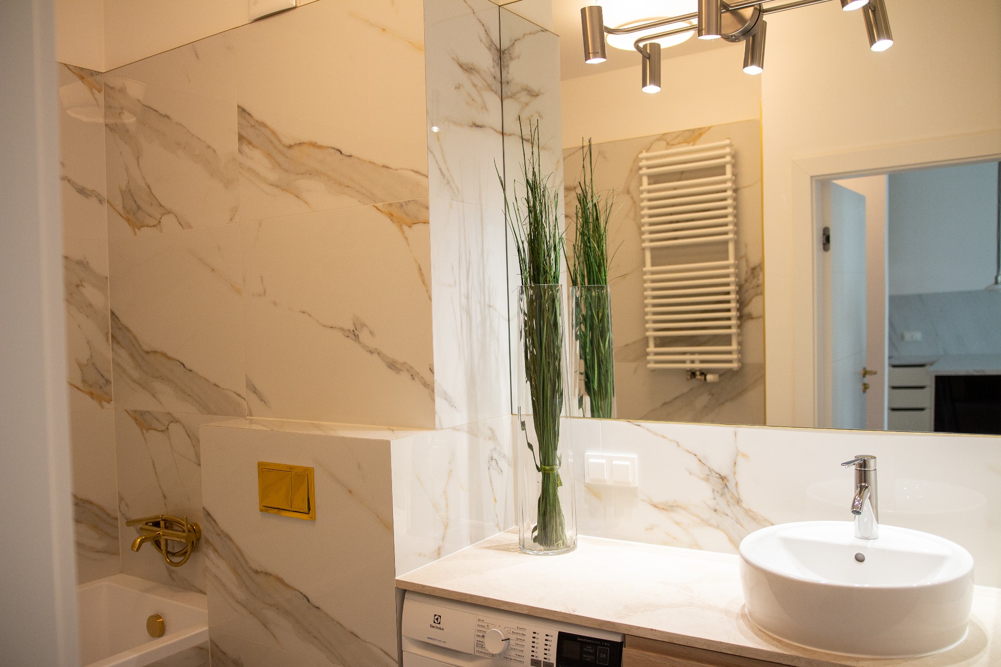 Elegancka łazienka z marmurowymi płytkami, złotą armaturą i umywalką nablatową, z pralką Electrolux w zabudowie pod blatem.