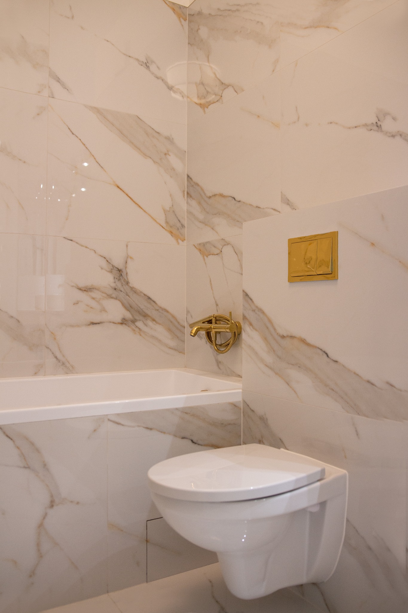 Wykończona łazienka z białymi płytkami imitującymi marmur, podwieszana toaleta i złota armatura.