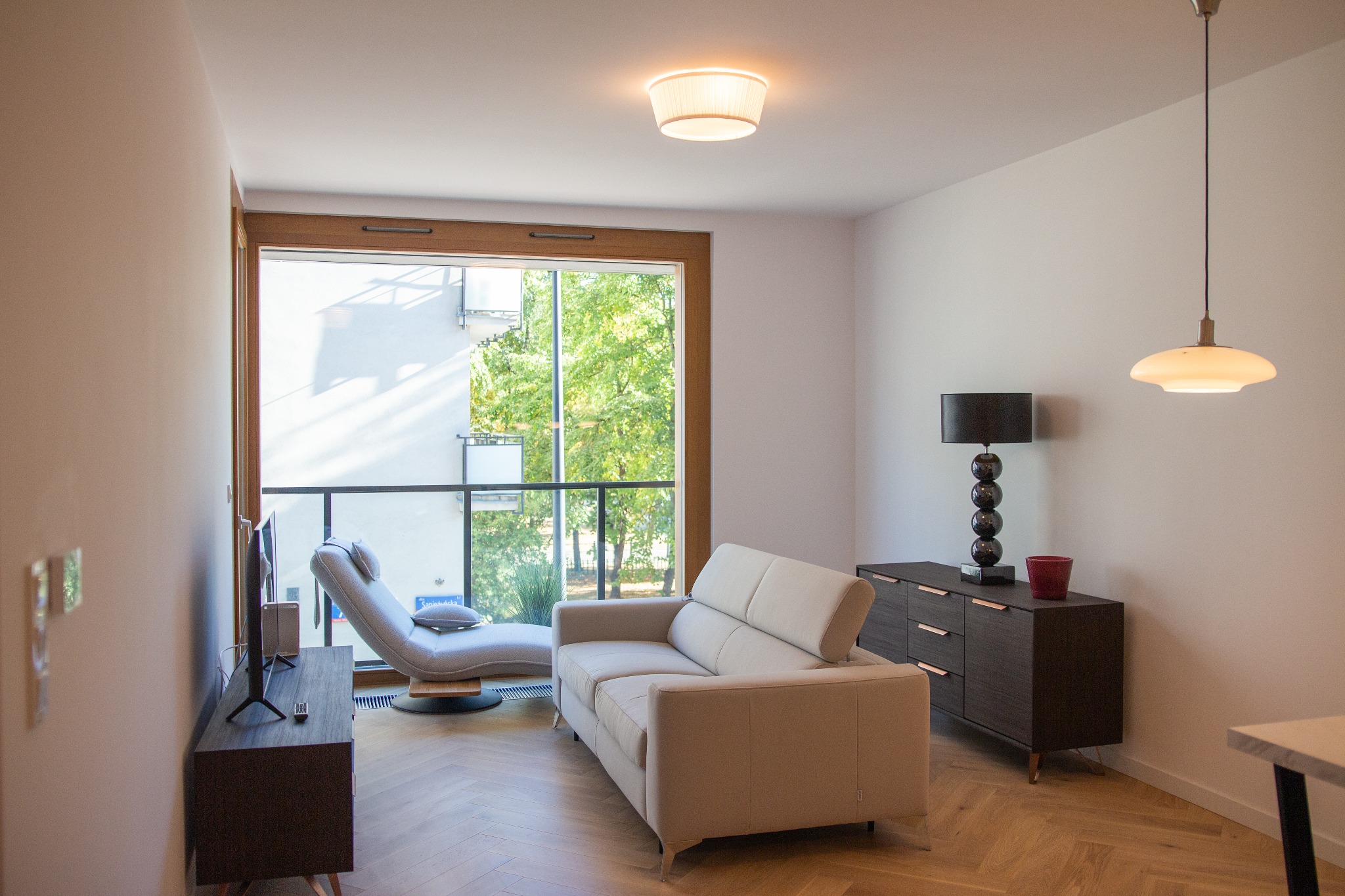 Jasne wnętrze salonu z parkietem w jodełkę, beżową sofą, szezlongiem przy balkonie i ciemną komodą z lampą kulkową.