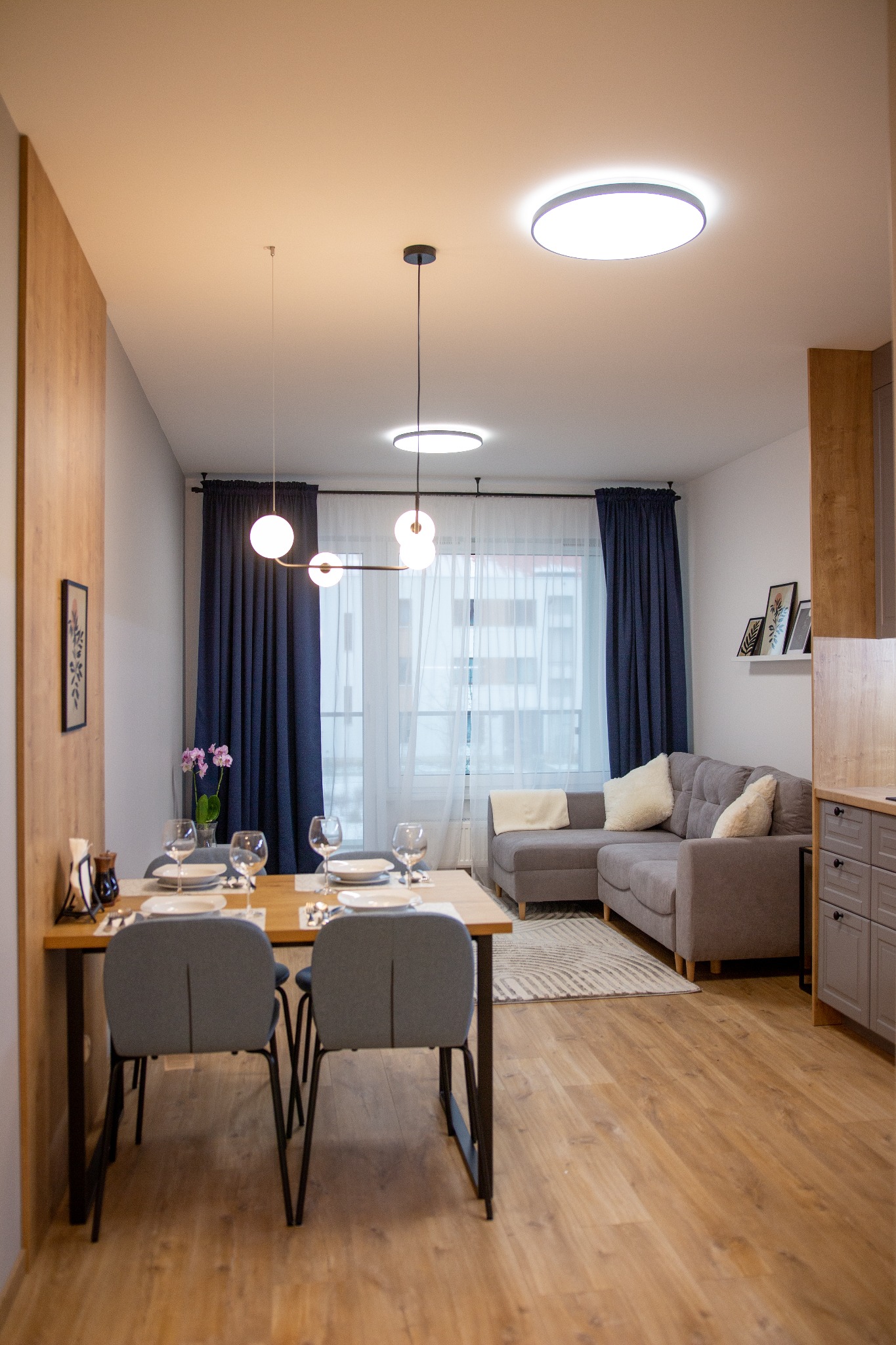 Jasne wnętrze mieszkania z drewnianą podłogą, szarą sofą narożną i stołem z nakryciem dla czterech osób, oświetlone nowoczesnymi lampami wiszącymi i sufitowymi.