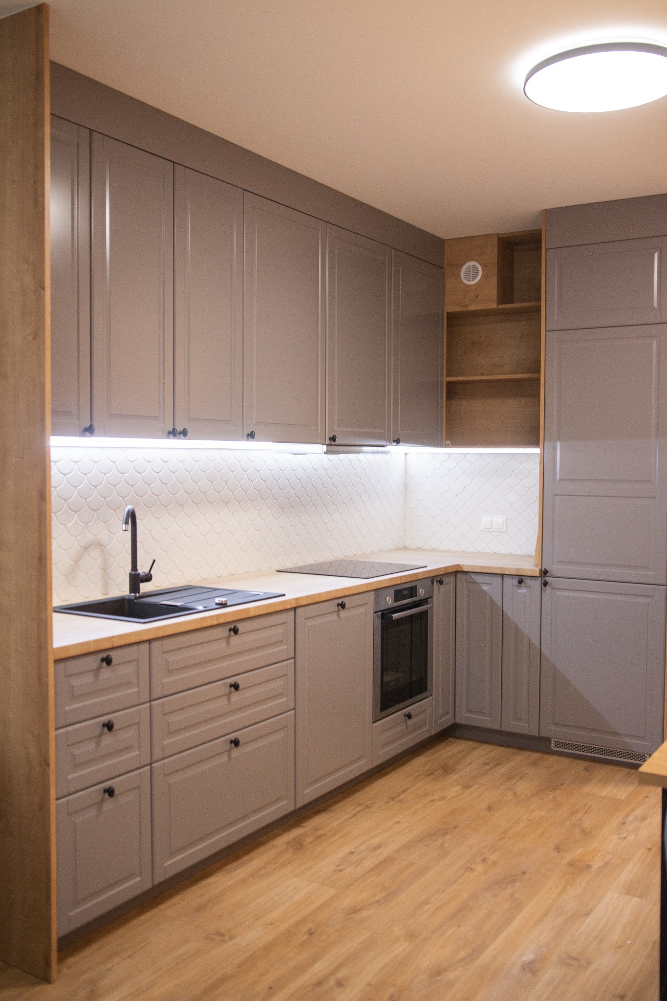 Szara, zabudowana kuchnia z drewnianym blatem, czarnym zlewem i baterią, oświetleniem LED pod szafkami oraz białymi płytkami w kształcie rybiej łuski.