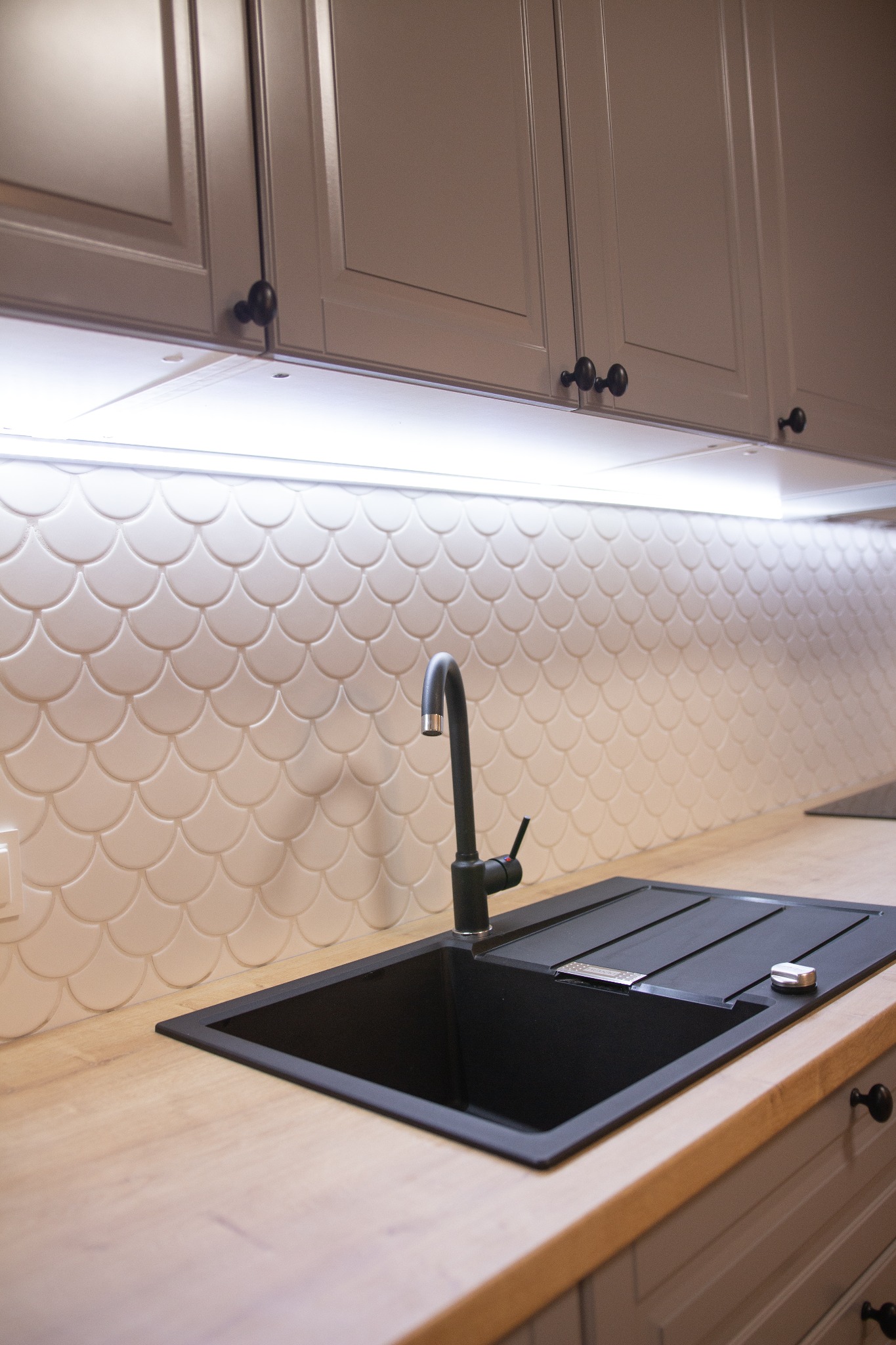 Nowoczesna kuchnia: czarny zlew wpuszczony w drewniany blat, bateria w kolorze czarnym, płytki w kształcie rybiej łuski na ścianie, oświetlenie LED pod szafkami górnymi, szare szafki z okrągłymi...