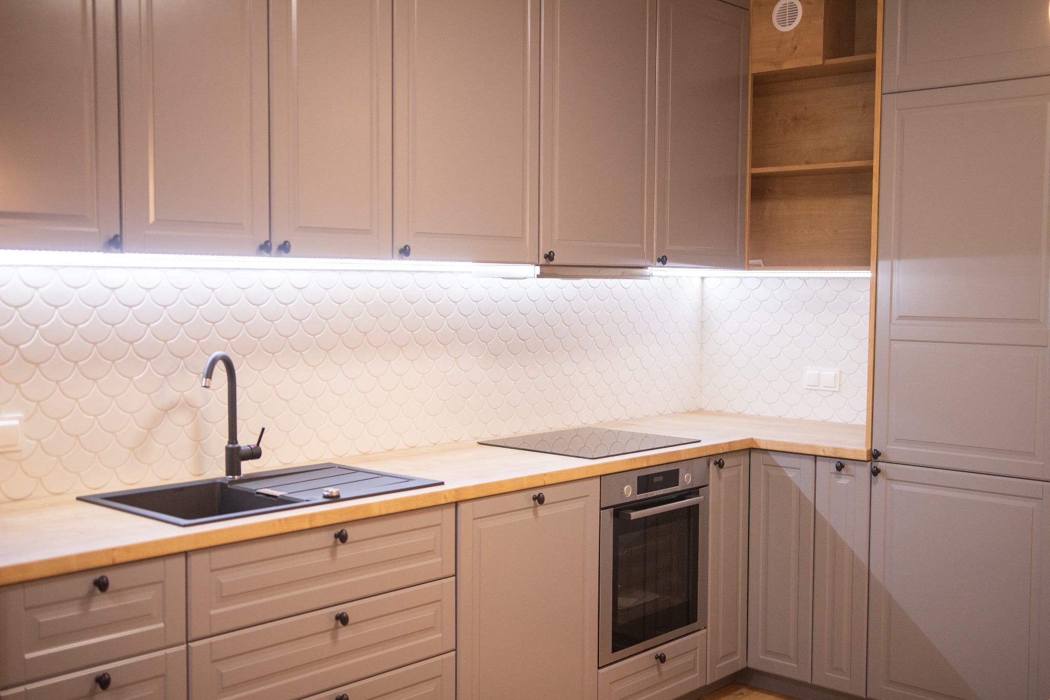 Szare meble kuchenne z drewnianym blatem i czarnym zlewem, wykończone pod klucz w mieszkaniu w Warszawie. Uwagę przykuwają płytki w kształcie rybiej łuski na ścianie.